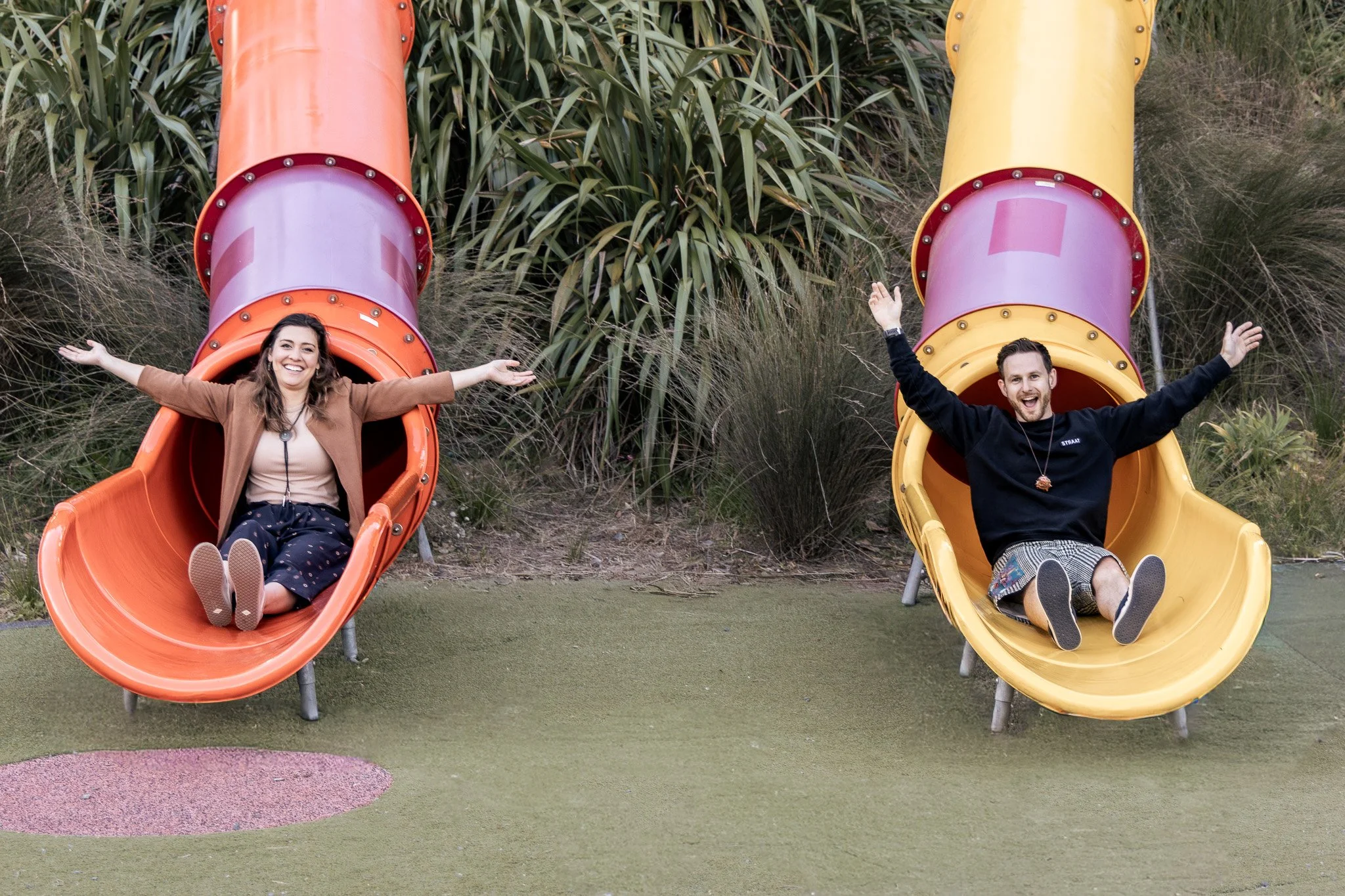 Two people, a woman and a man, are sliding down colorful playground slides outdoors surrounded by greenery and bushes. They are smiling and have their arms open wide.