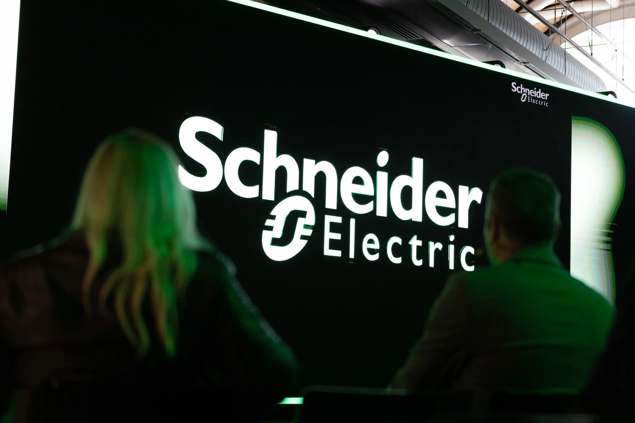 Close-up of illuminated neon sign reading 'Schneider Electric' in a modern, industrial setting with blurred figures in the foreground.