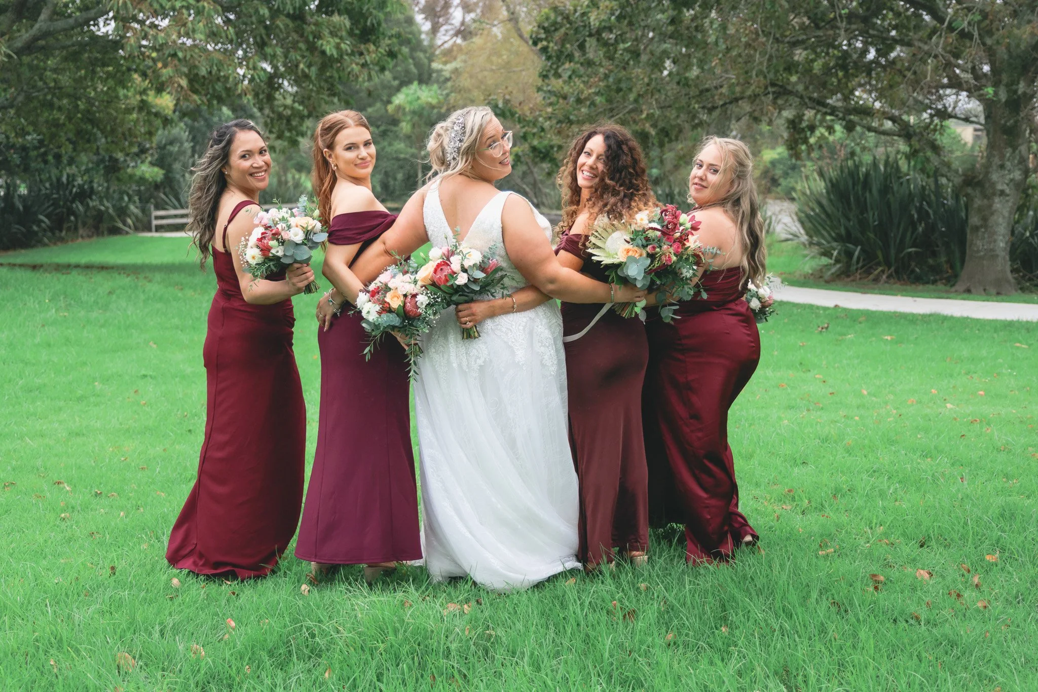 A group of five women, one in a white wedding dress and four in maroon bridesmaid dresses, smiling and holding bouquets, standing on a grassy area outdoors surrounded by trees.