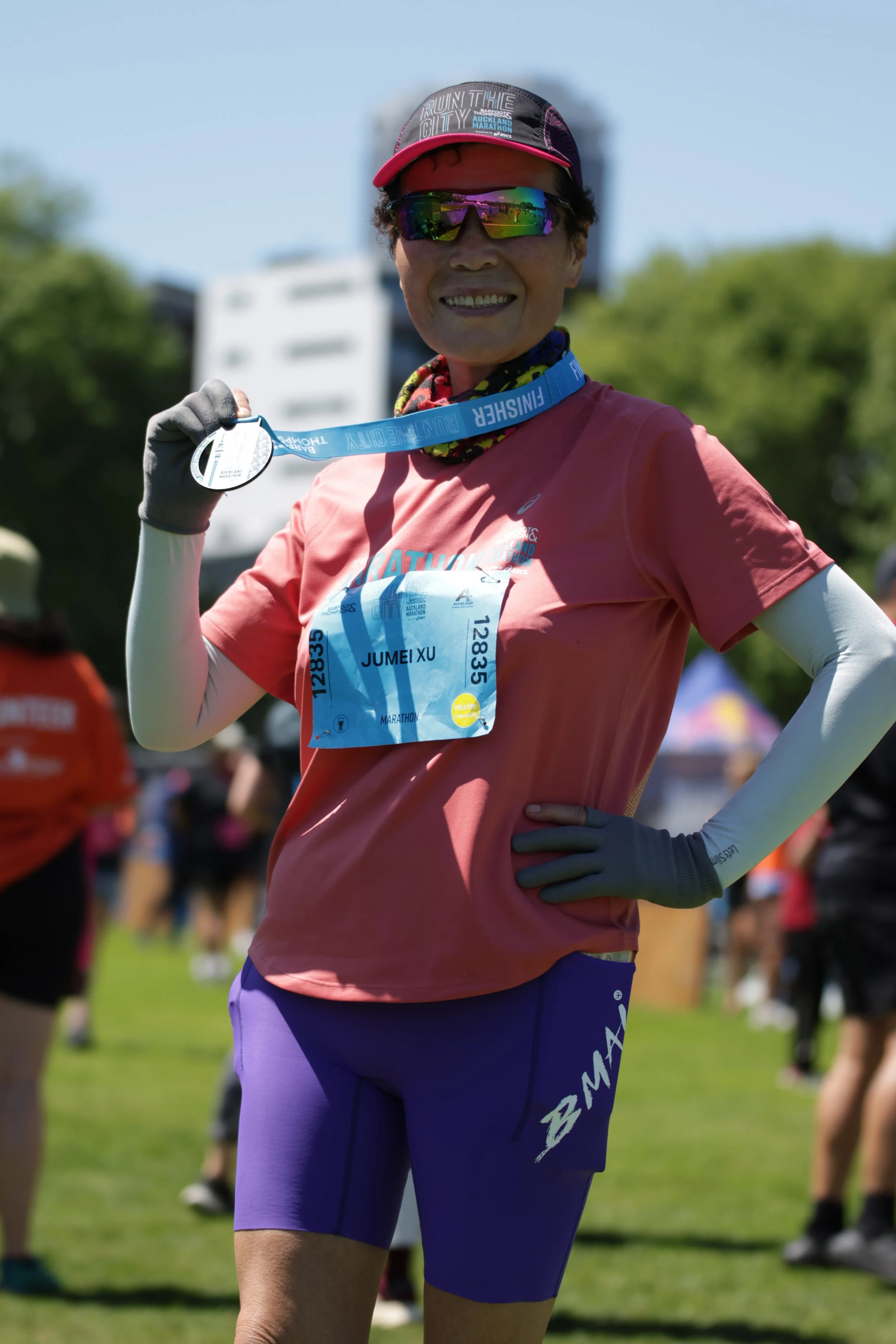 A smiling woman wearing sunglasses and medals around her neck, posing outdoors at a marathon event, with other runners in the background.