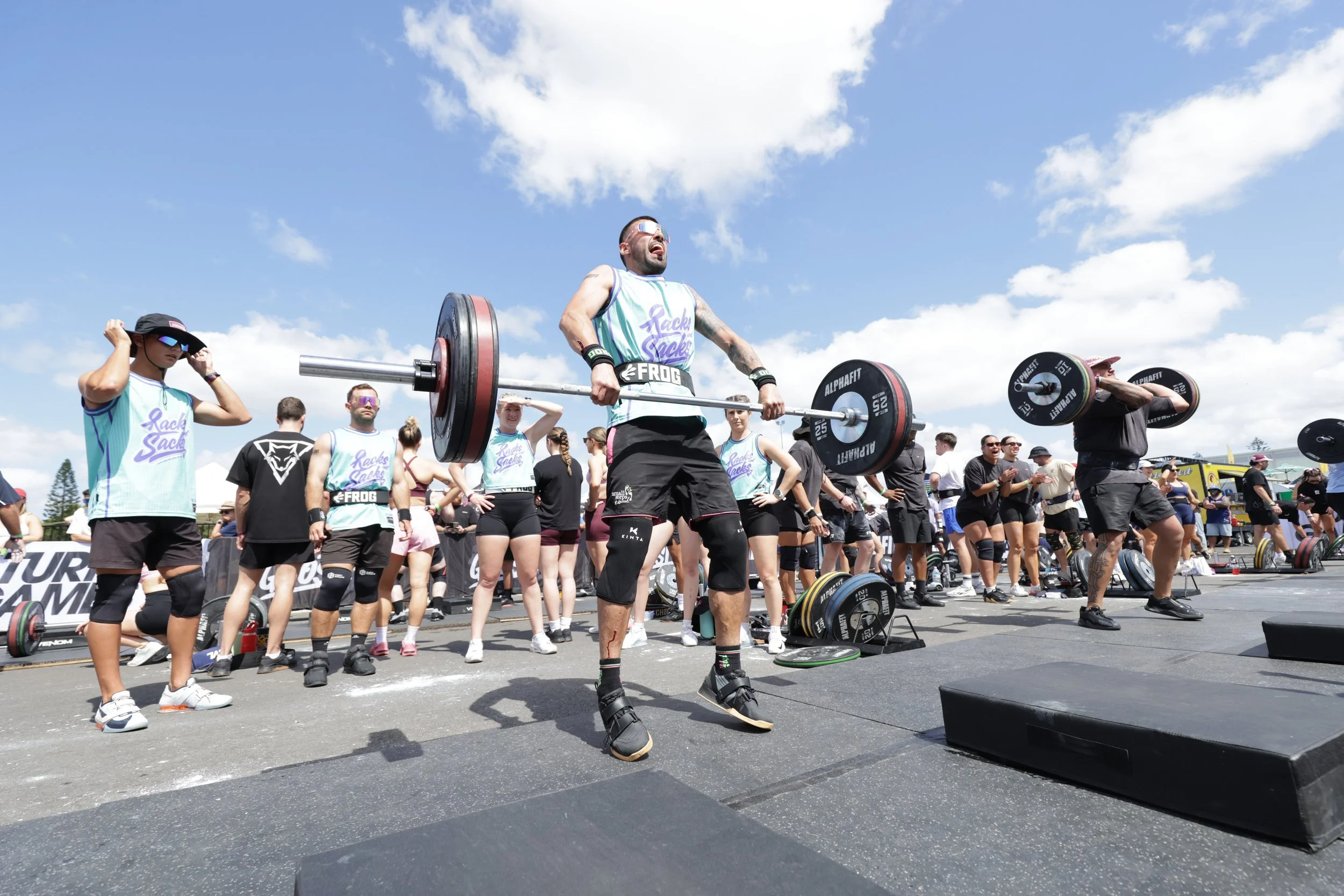 A group of people at an outdoor weightlifting event, with some lifting barbells and others observing, under a blue sky with clouds.