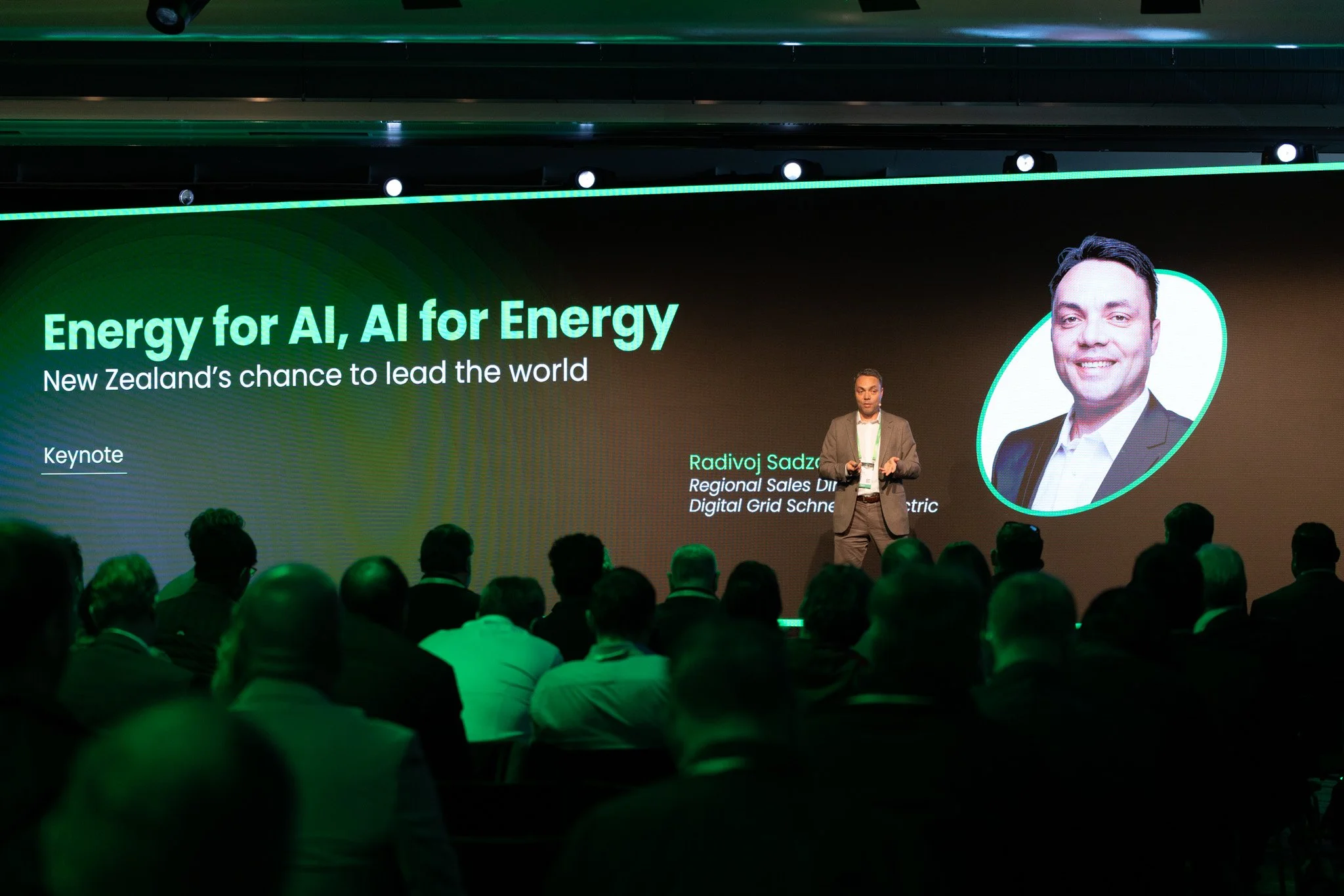 A man in a gray suit giving a presentation on a stage in front of an audience, with a large screen displaying the words 'Energy for AI, AI for Energy' and a photo of another man. The event appears to be a conference or keynote.