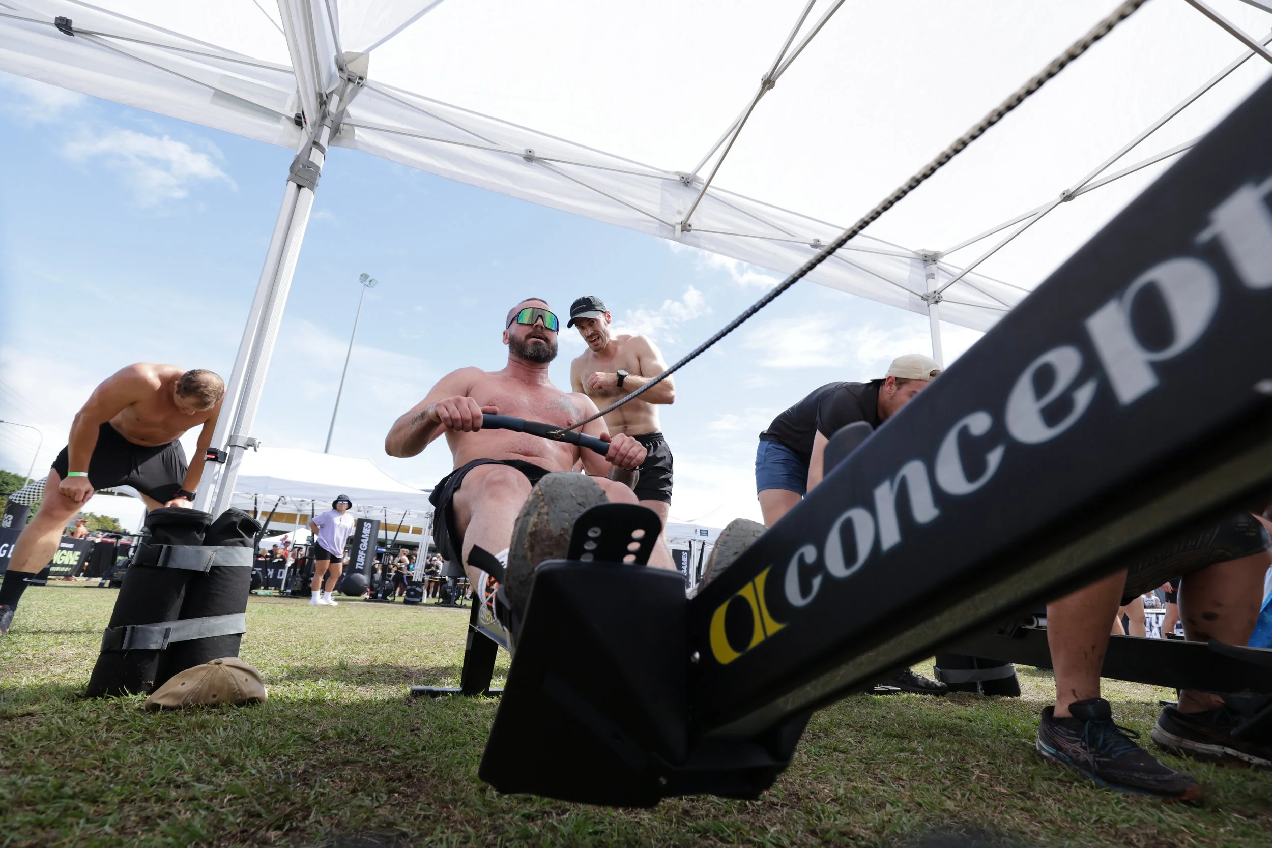 Muscular man with sunglasses and a tattoo on his chest participating in a strongman competition, pulling a heavy sled with multiple weights, under a white canopy at an outdoor event with other athletes and spectators nearby.