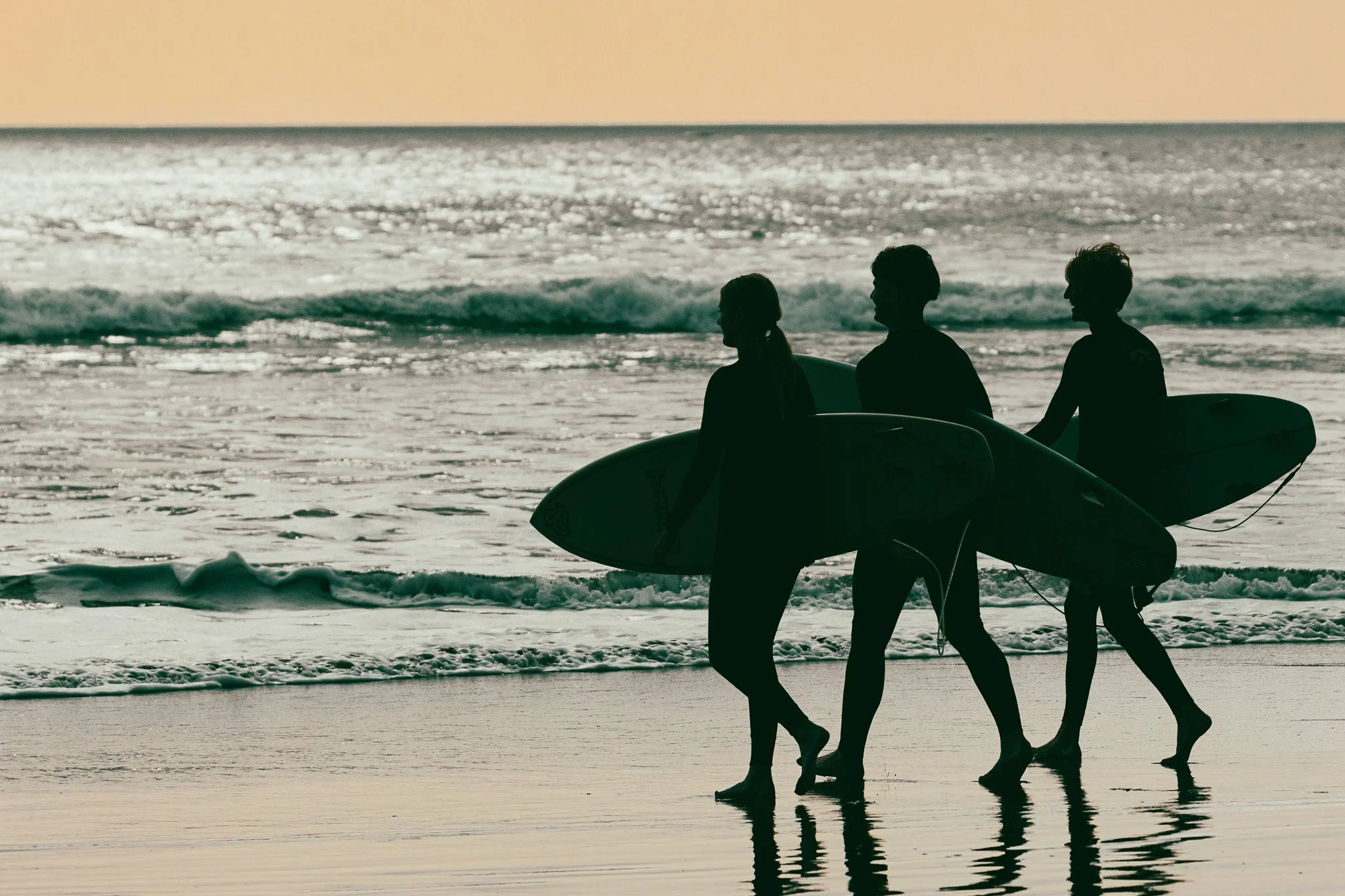 Silhouettes of three surfers walking along the beach with surfboards, near the shore during sunset or sunrise, with gentle waves in the background.