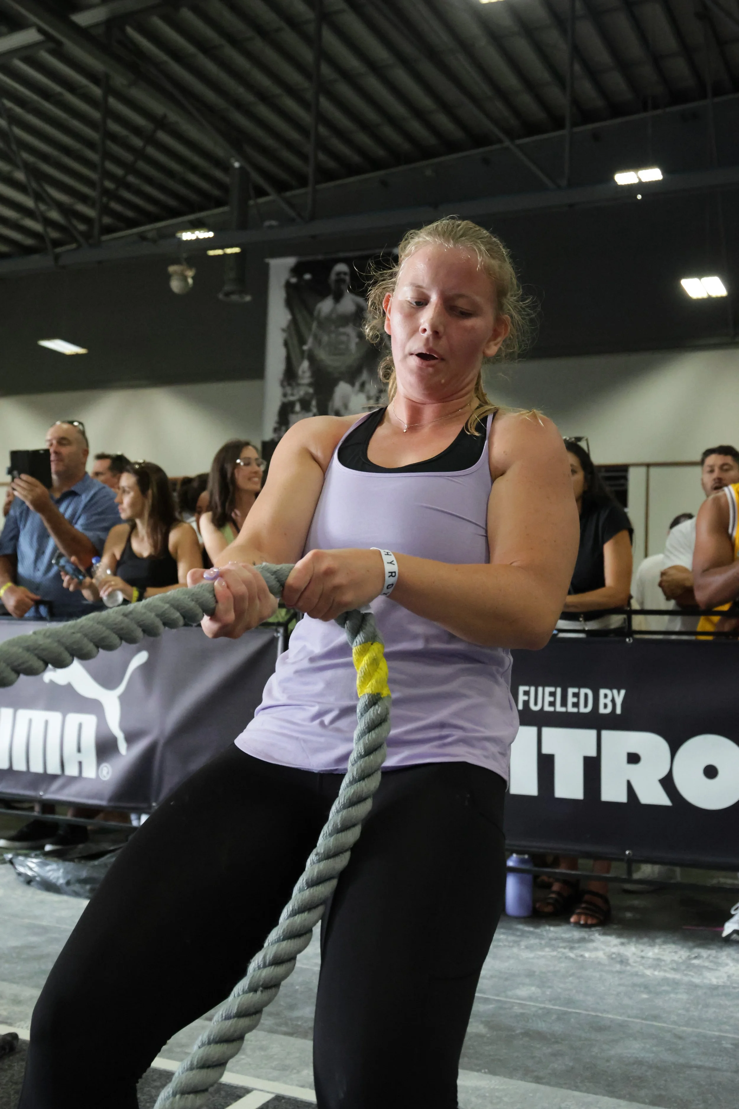 A woman in athletic clothing participating in a tug-of-war competition at an indoor event with a crowd watching.