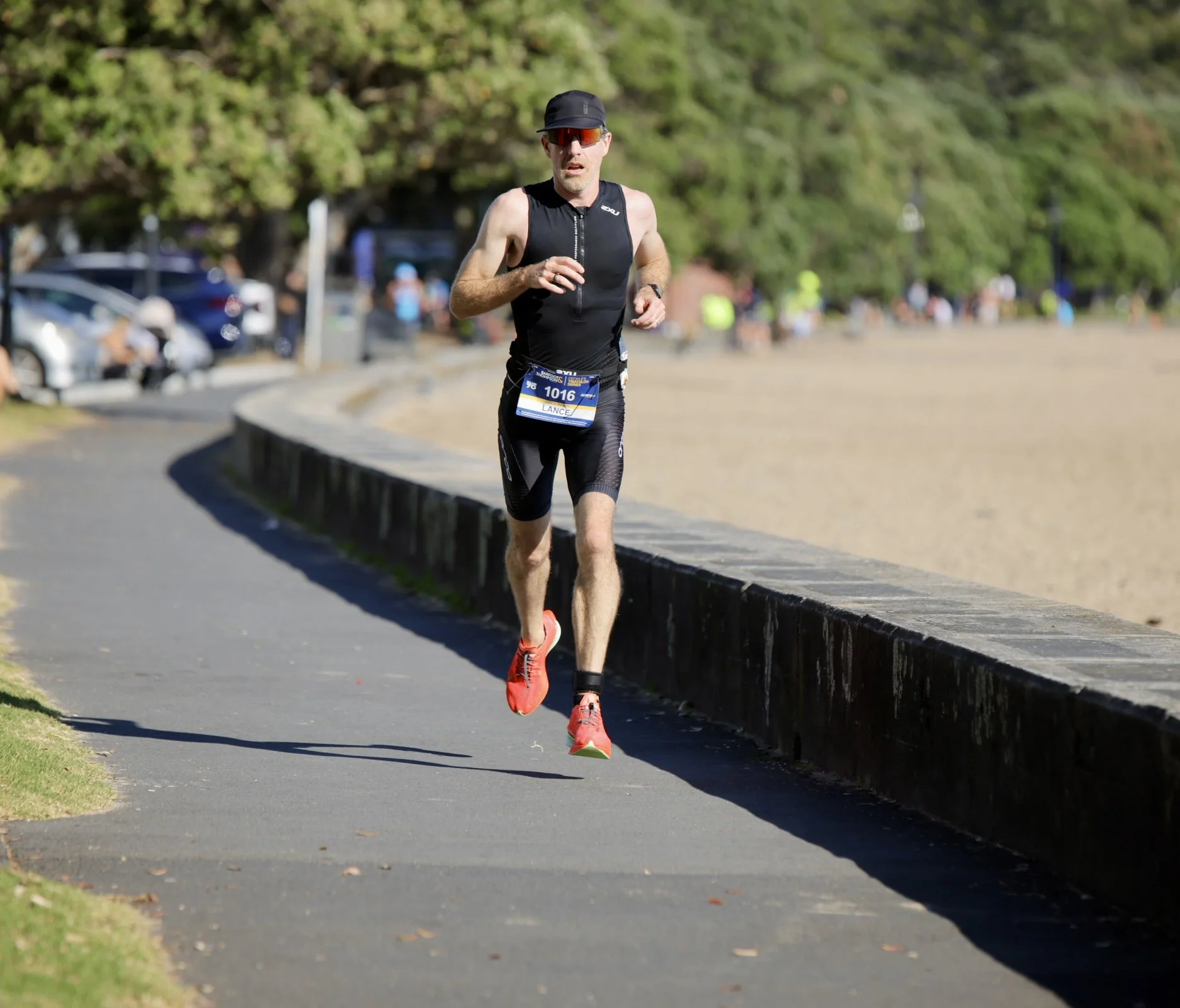 A male runner in black athletic gear running along a paved path with a low stone wall on his right, in a park with green trees and blurry spectators in the background.