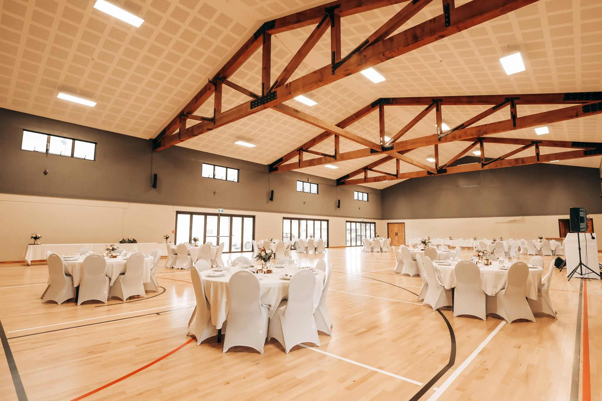 Large indoor event space set up for a banquet with round tables covered in white tablecloths and chairs with white covers, decorated with floral centerpieces. The ceiling features exposed wooden beams and bright lighting. Windows are along the upper 