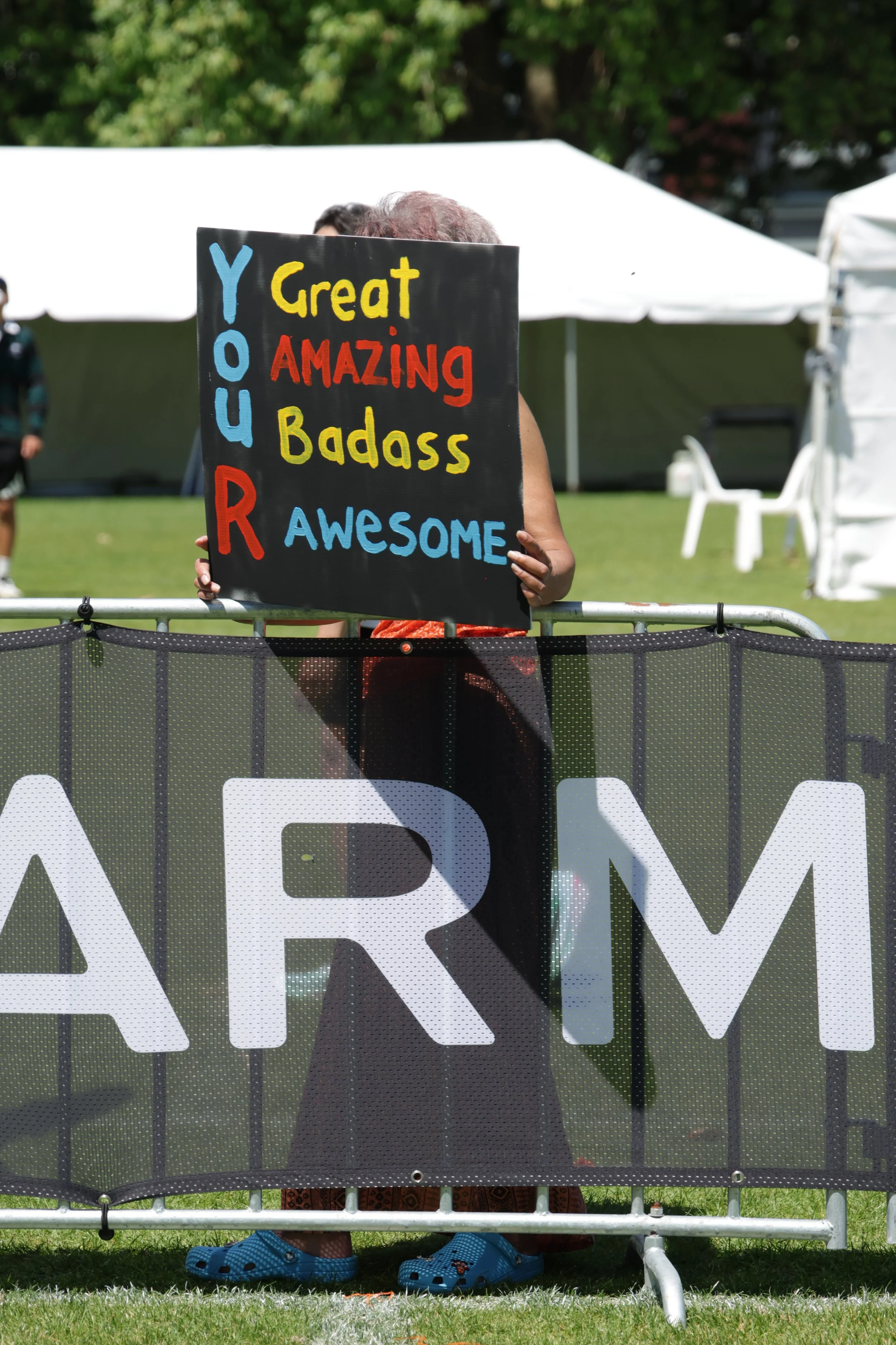 Person kneeling behind a black sign with colorful words using a barrier for coverage, with white tents and green trees in the background.