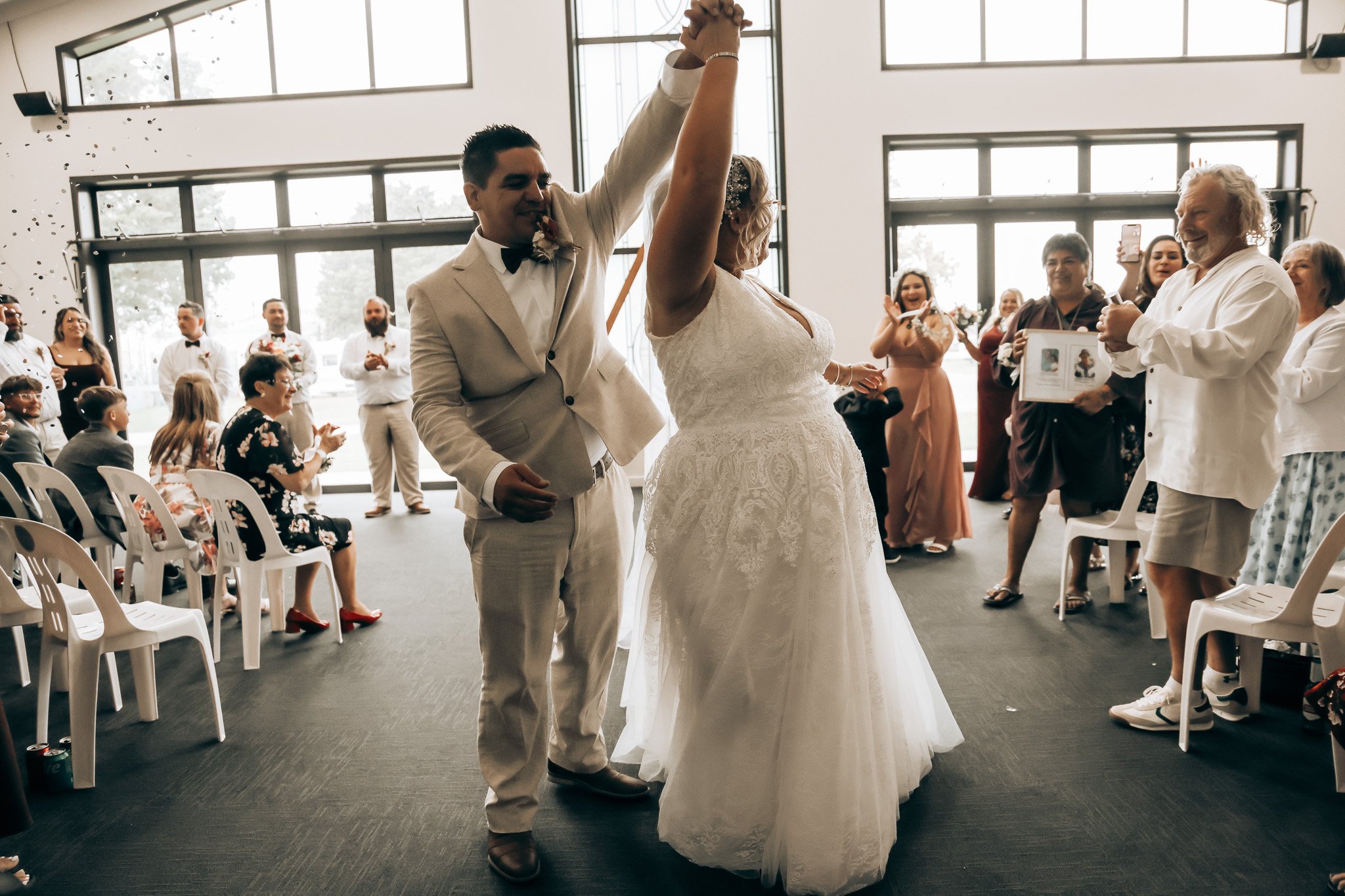 A bride and groom dancing at their wedding reception with guests smiling and clapping around them inside a large room with lots of windows.
