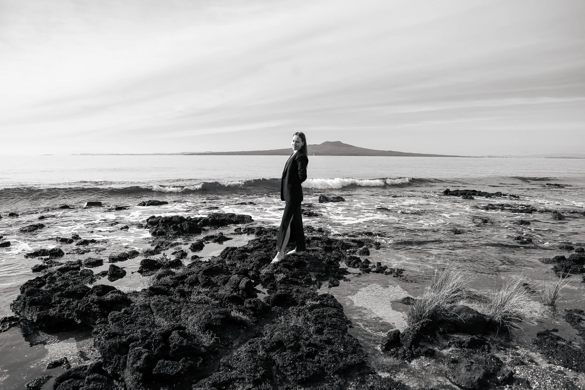 A woman in formal attire standing on rocks at the edge of the ocean with a body of water and island in the background.
