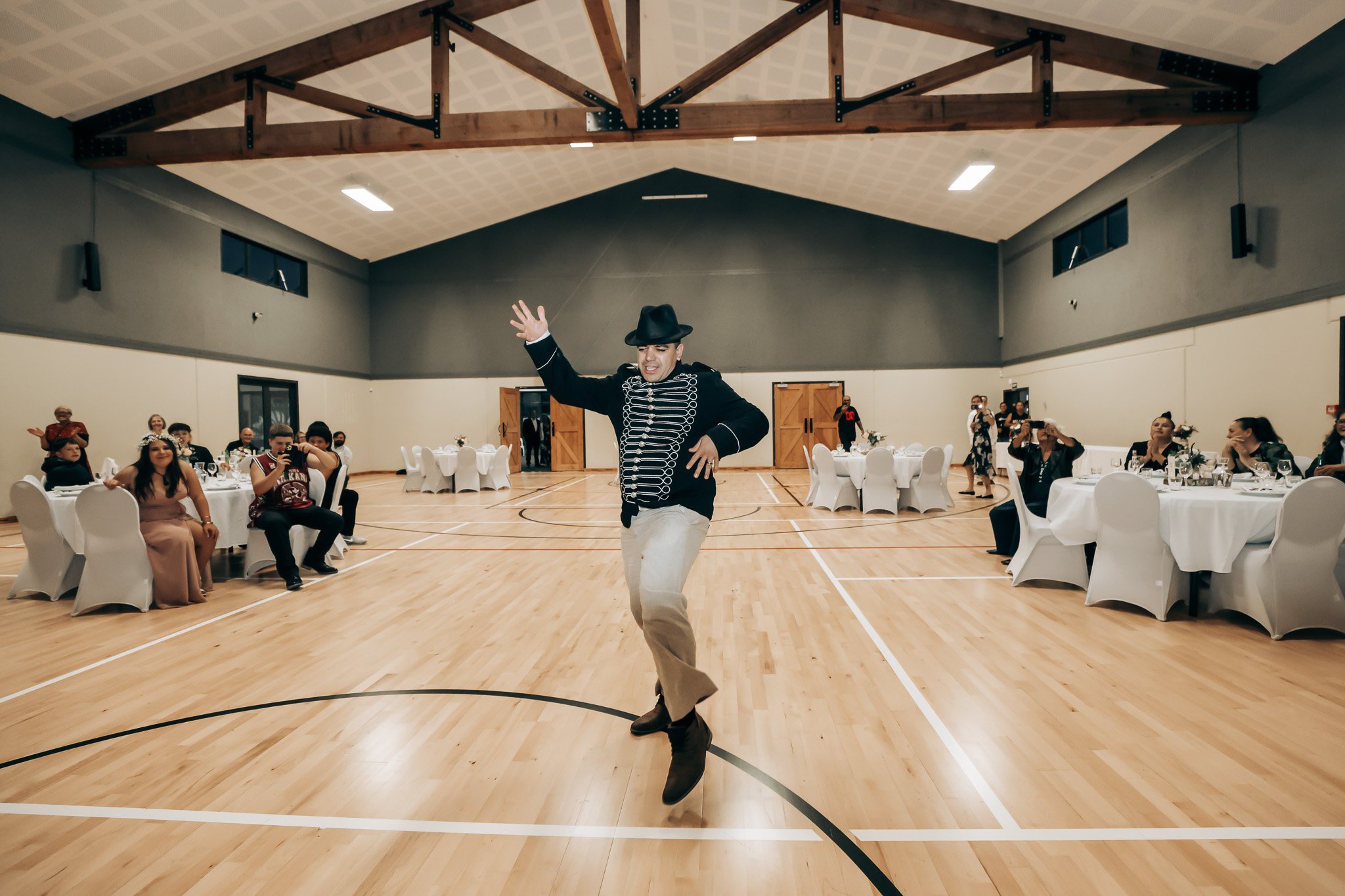 A man dressed in a black jacket with white piping, beige pants, and wearing a black hat is dancing in a gymnasium with a wooden floor. He is in the center of the image, with his left arm raised and his right hand near his waist, and appears to be enj