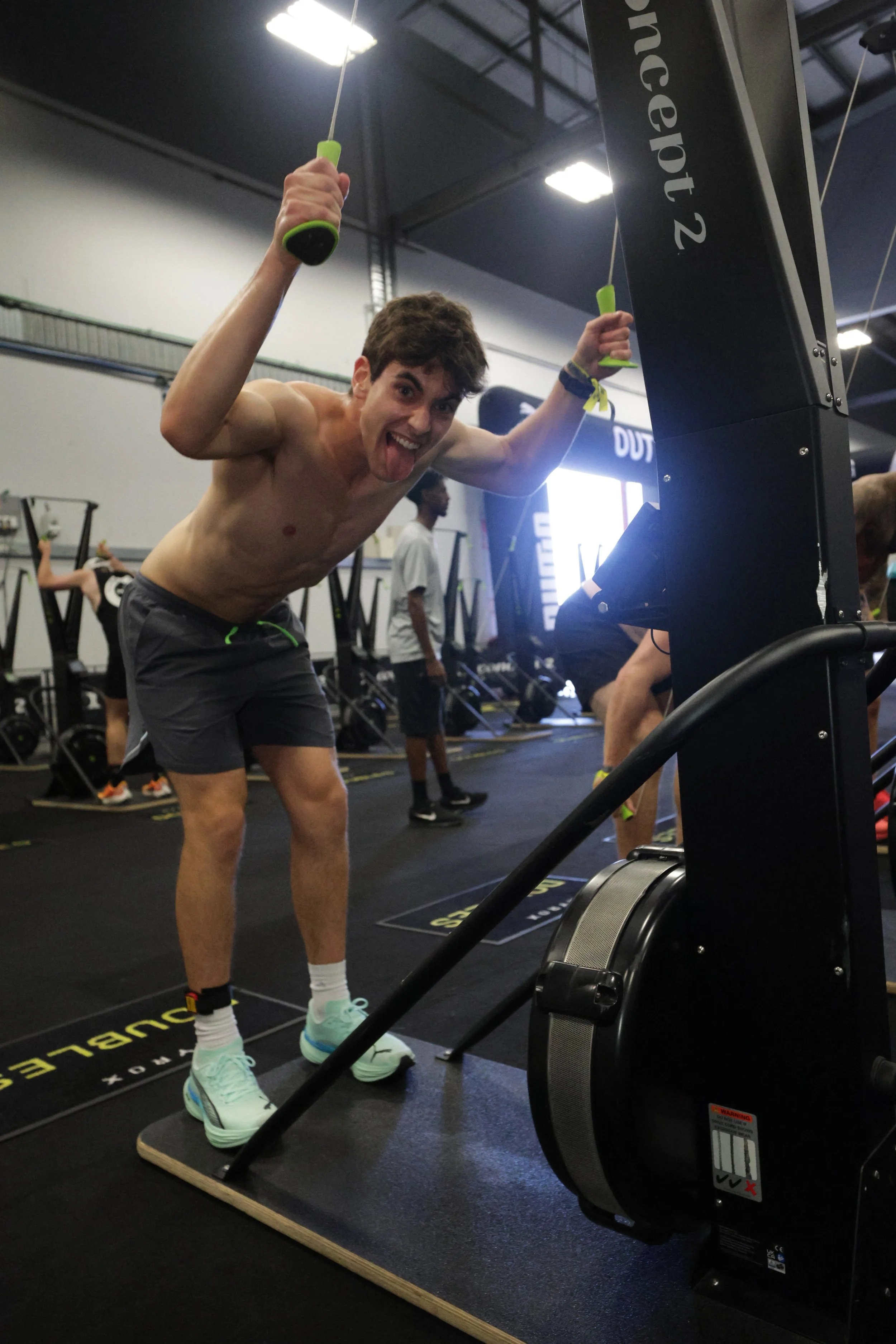 A shirtless man with a lollipop sticking out of his mouth and a playful expression is standing on a wooden balance board at a gym, holding onto the handles of a piece of gym equipment. The gym has black flooring, high ceilings, and various exercise m