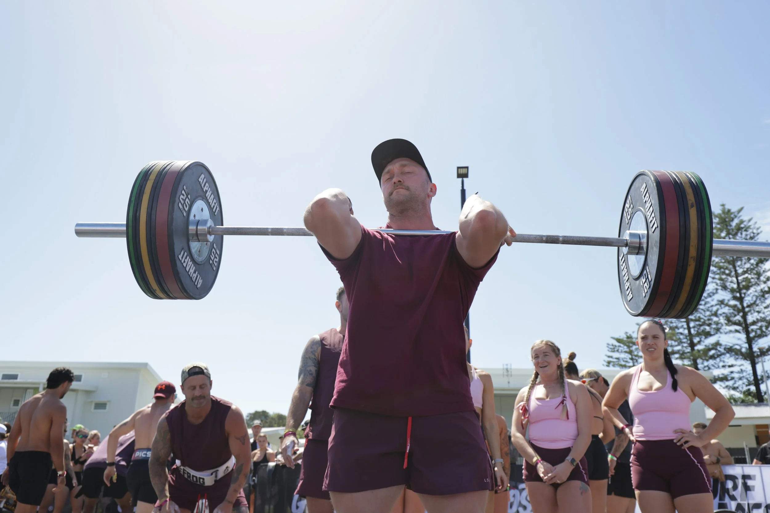 A man in a maroon shirt and black cap lifting a loaded Olympic barbell during a weightlifting event outdoors, surrounded by other athletes and spectators.