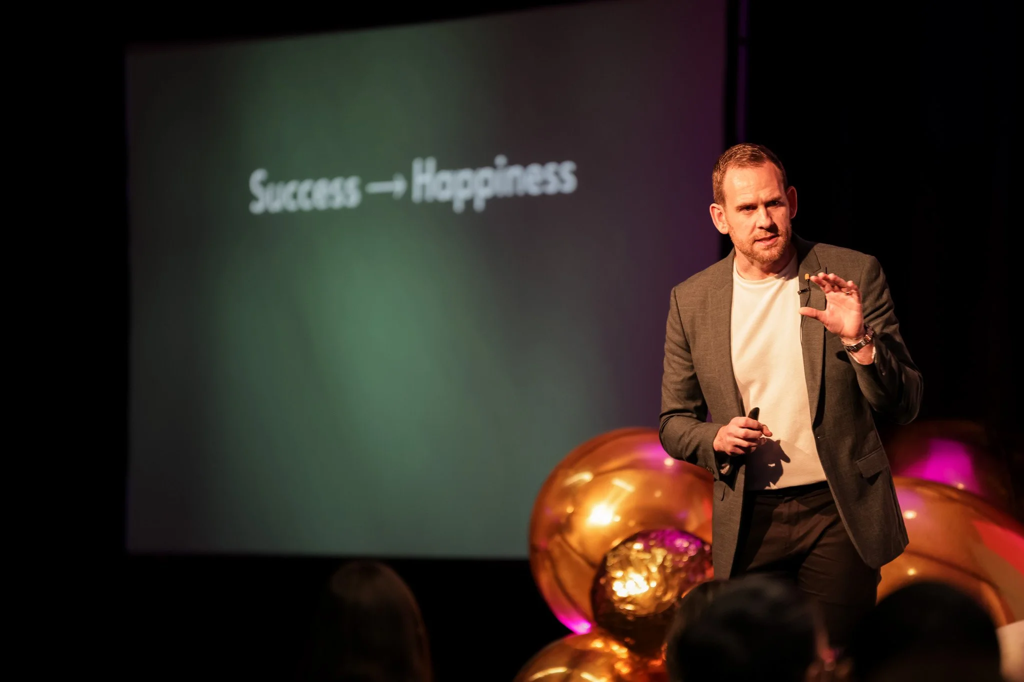 A man in a gray blazer and white shirt giving a presentation on stage with a dark background and a slide that reads 'Success → Happiness'. There are gold balloons in the foreground.