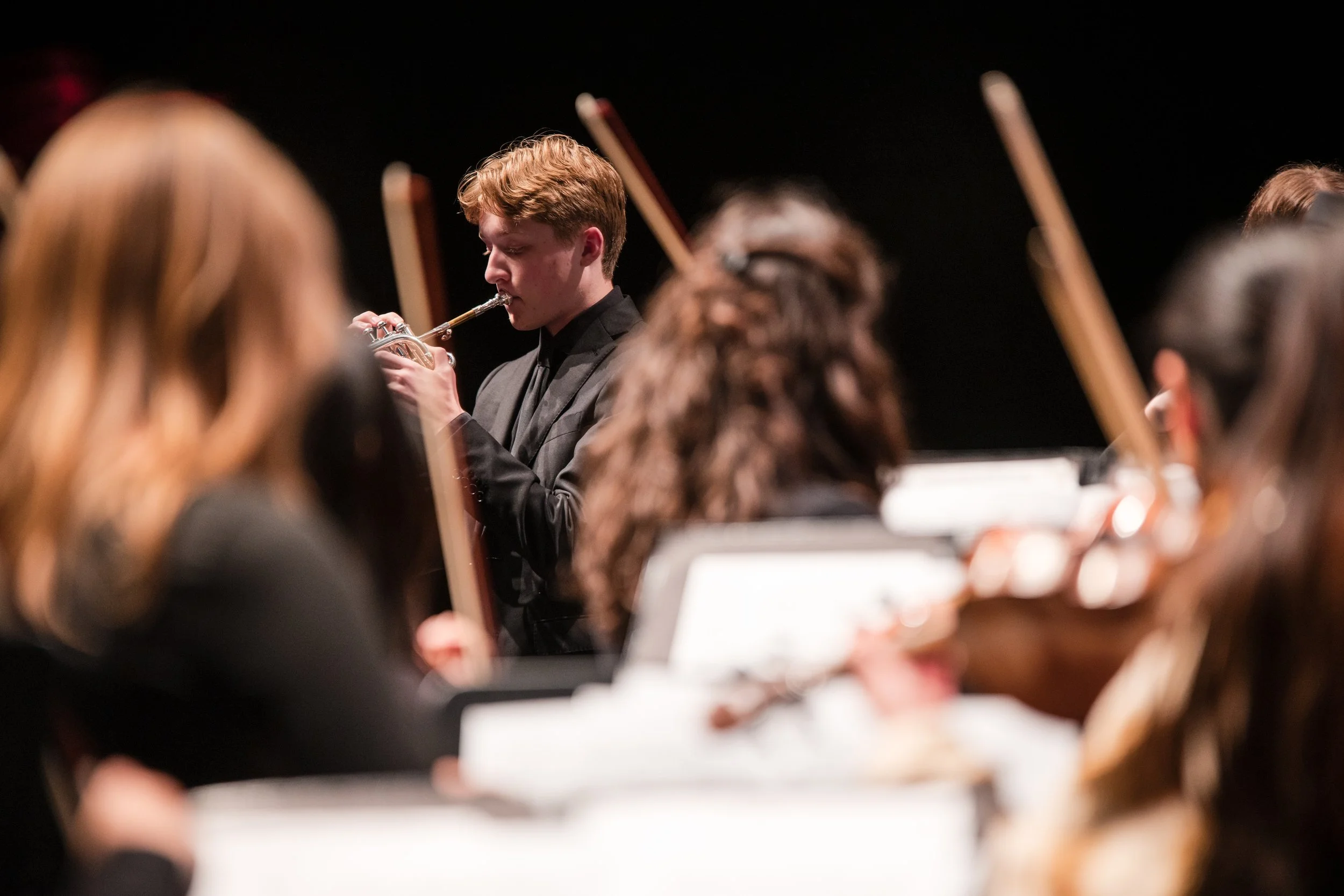 Young man playing a trumpet in an orchestra, with other musicians around him, during a concert.