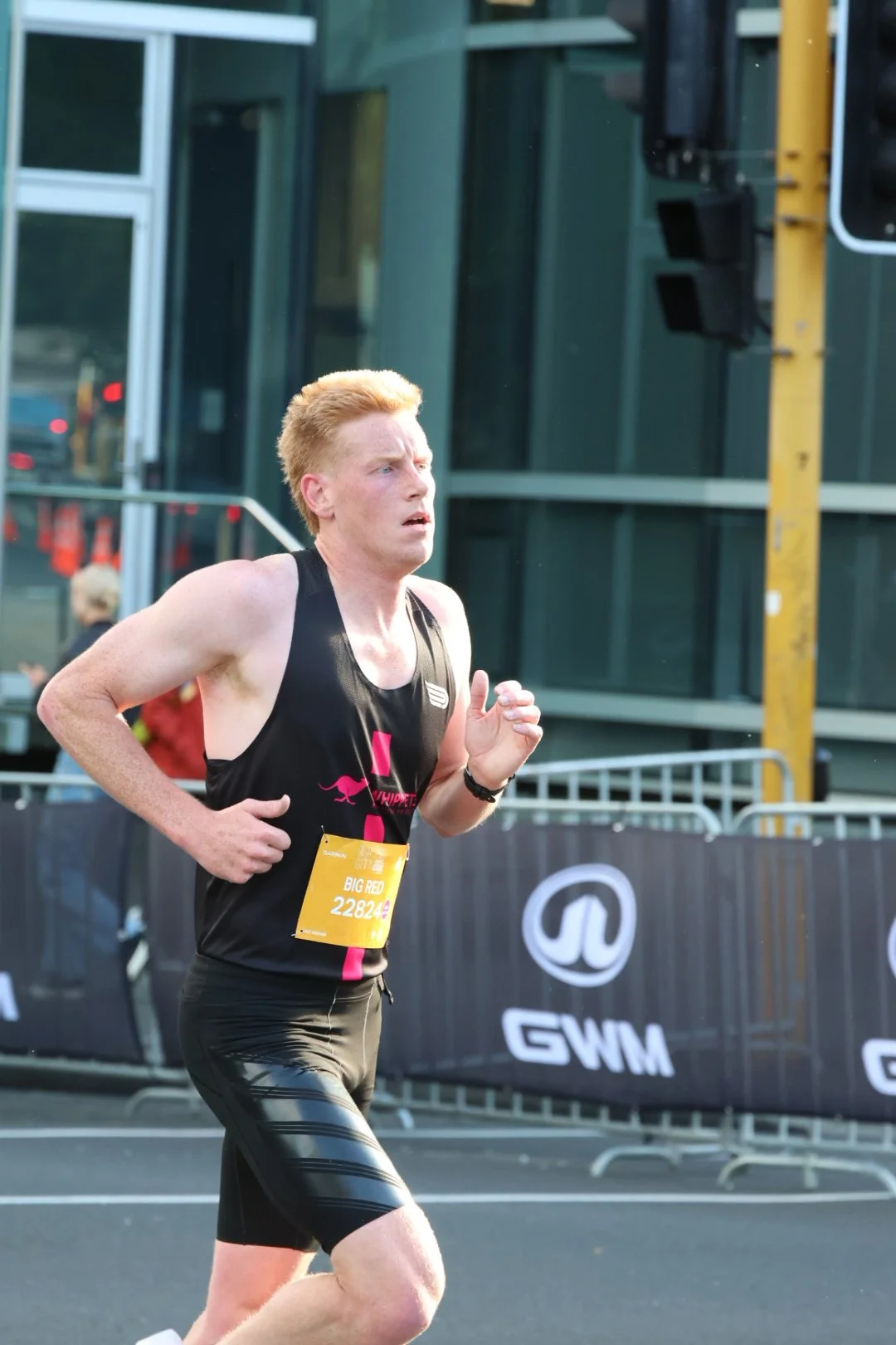 A male marathon runner with red hair and wearing a black athletic outfit running in an urban setting during daylight.