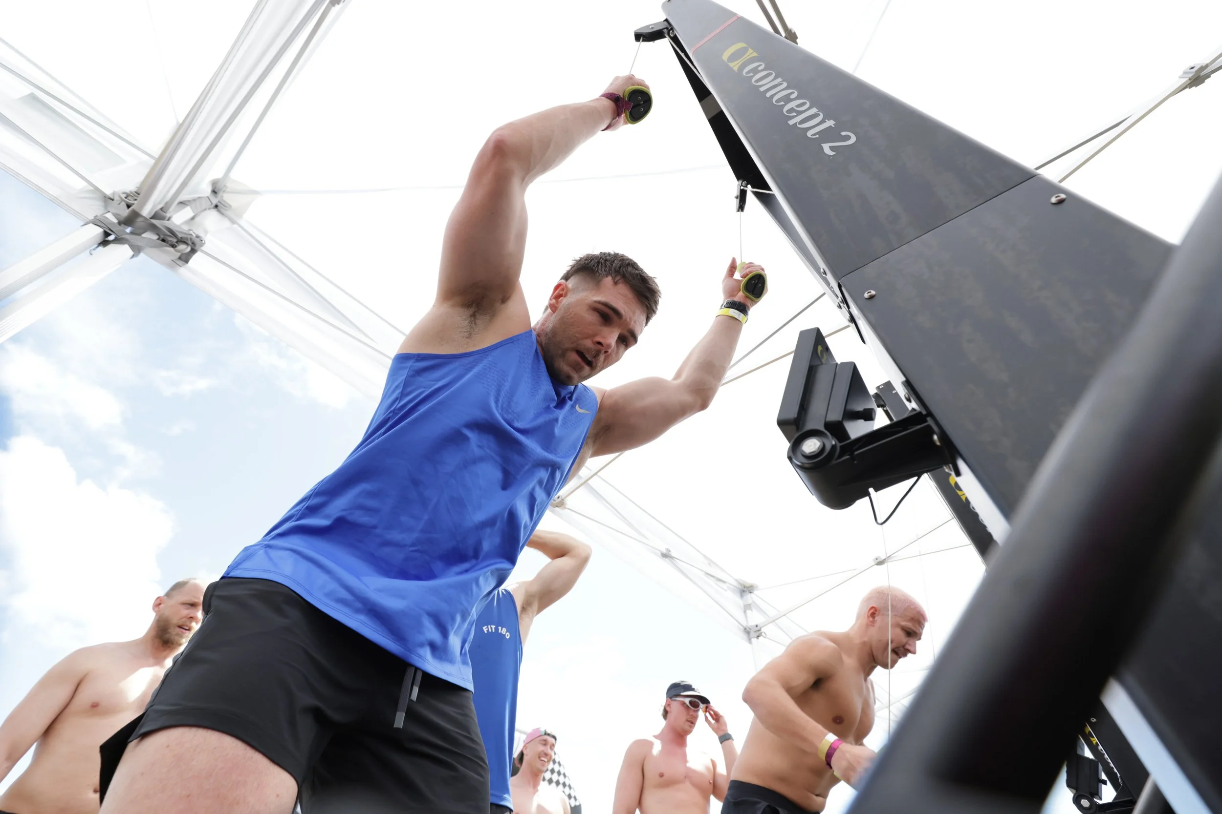 A man in a blue shirt preparing for a rowing event with several shirtless men in the background, under a tent on a cloudy day.