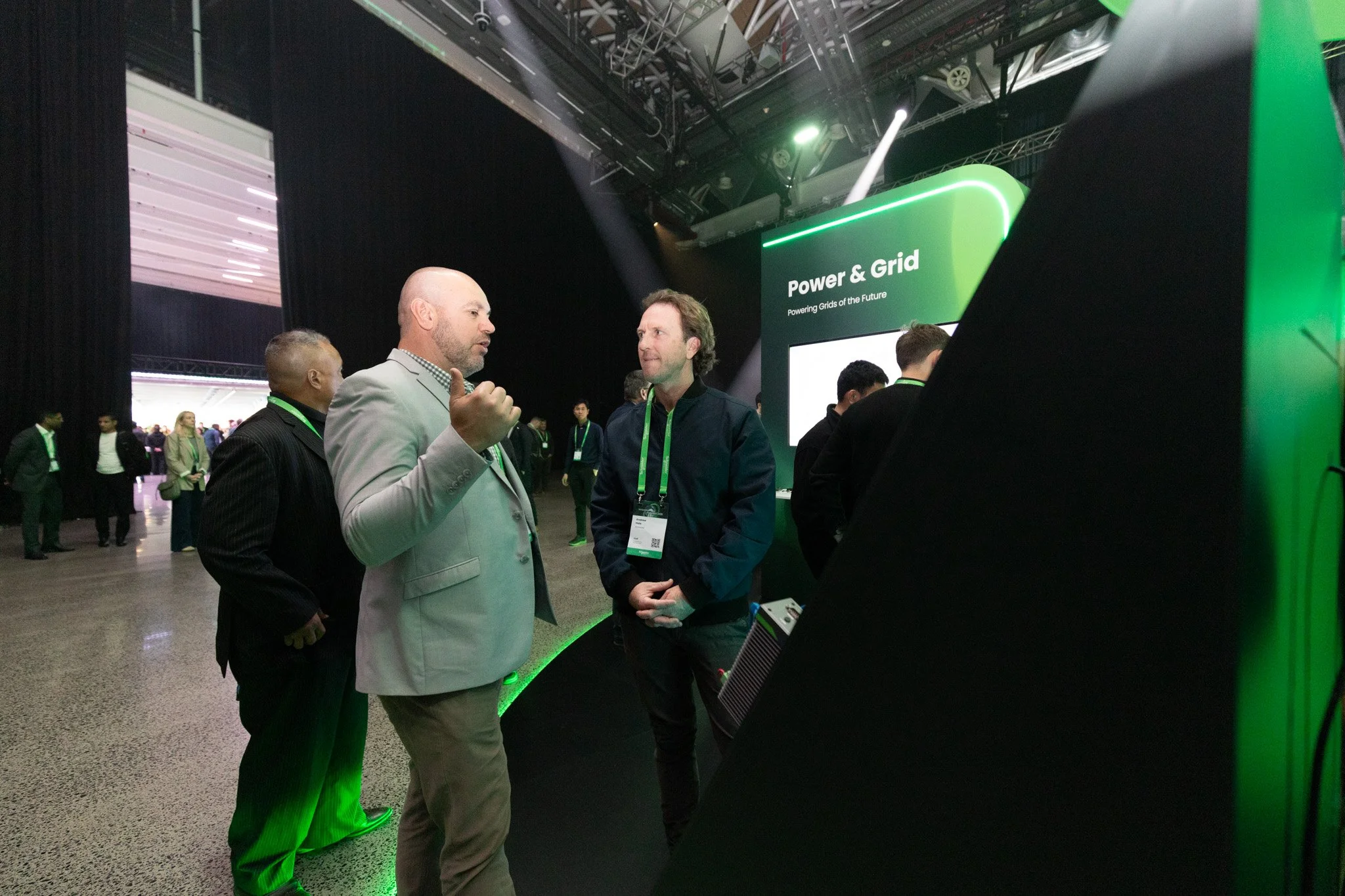 Three men are engaged in conversation at a technology conference called Power & Grid, with a sign behind them reading 'Power & Grid: Powering Grids of the Future.' The venue has a high ceiling and multiple attendees in the background.