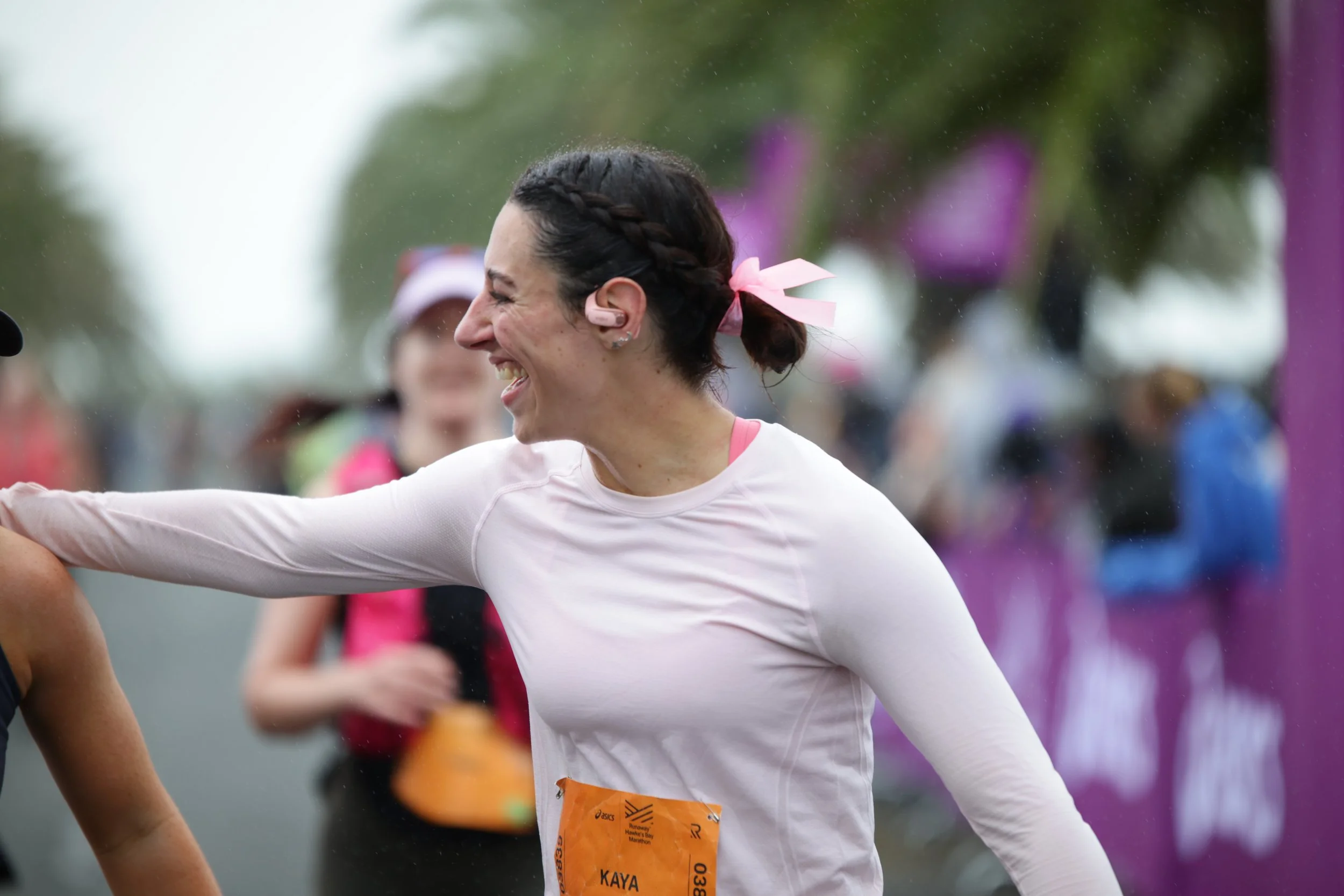A woman with dark hair braided, wearing a white long-sleeve shirt and pink ribbon in her hair, smiling and participating in a marathon with a bib tag that reads Kay.