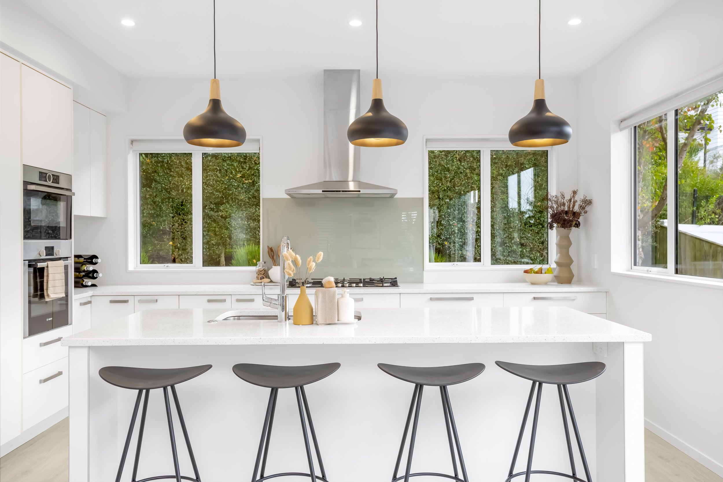 Modern white kitchen with black pendant lights, a kitchen island with bar stools, and large windows showing greenery outside.