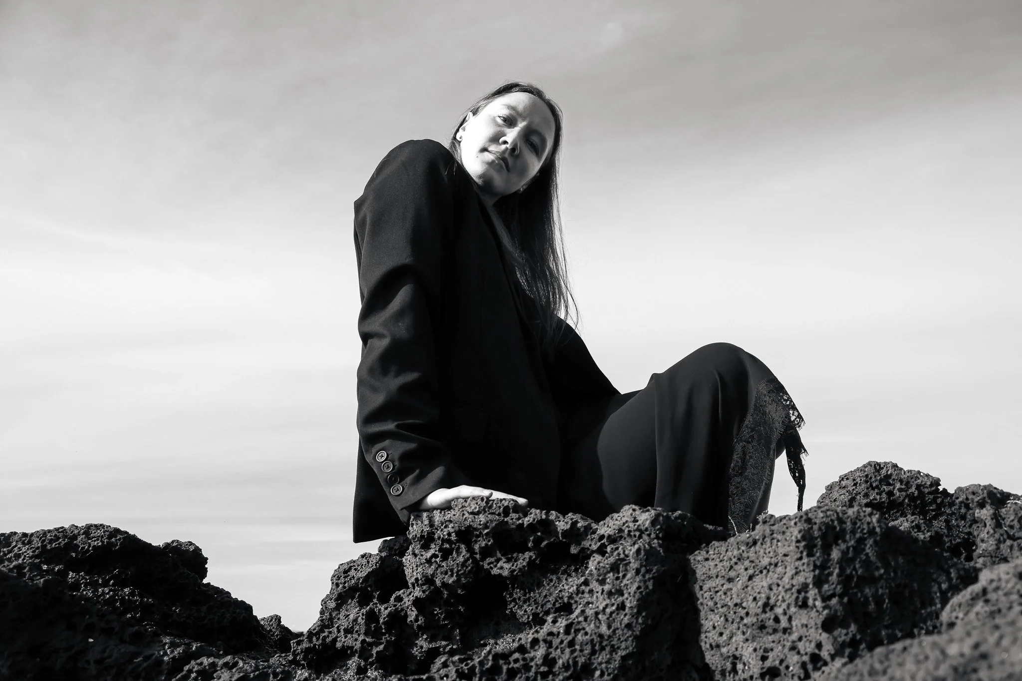 Black and white photo of a woman sitting on dark volcanic rocks, looking at the camera with a serious expression, wearing a dark blazer, outdoors against a cloudy sky.