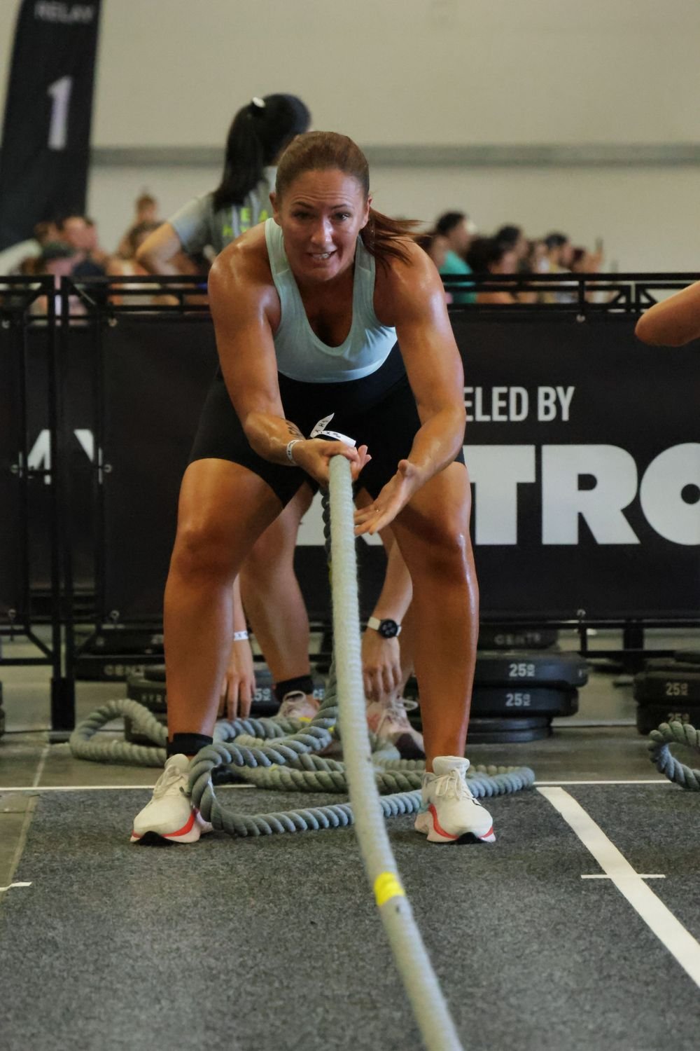 A woman participating in a strongman competition, pulling a rope with focused effort. She is wearing athletic clothing and sneakers, with a crowd of spectators and a large banner in the background.
