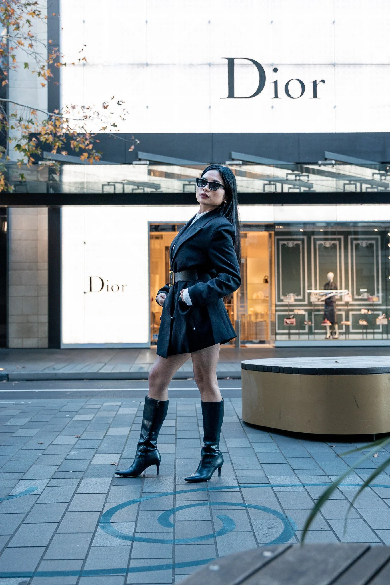 A woman standing on a city sidewalk wearing a black blazer dress and high-heeled leather boots, with sunglasses, in front of a Dior storefront.