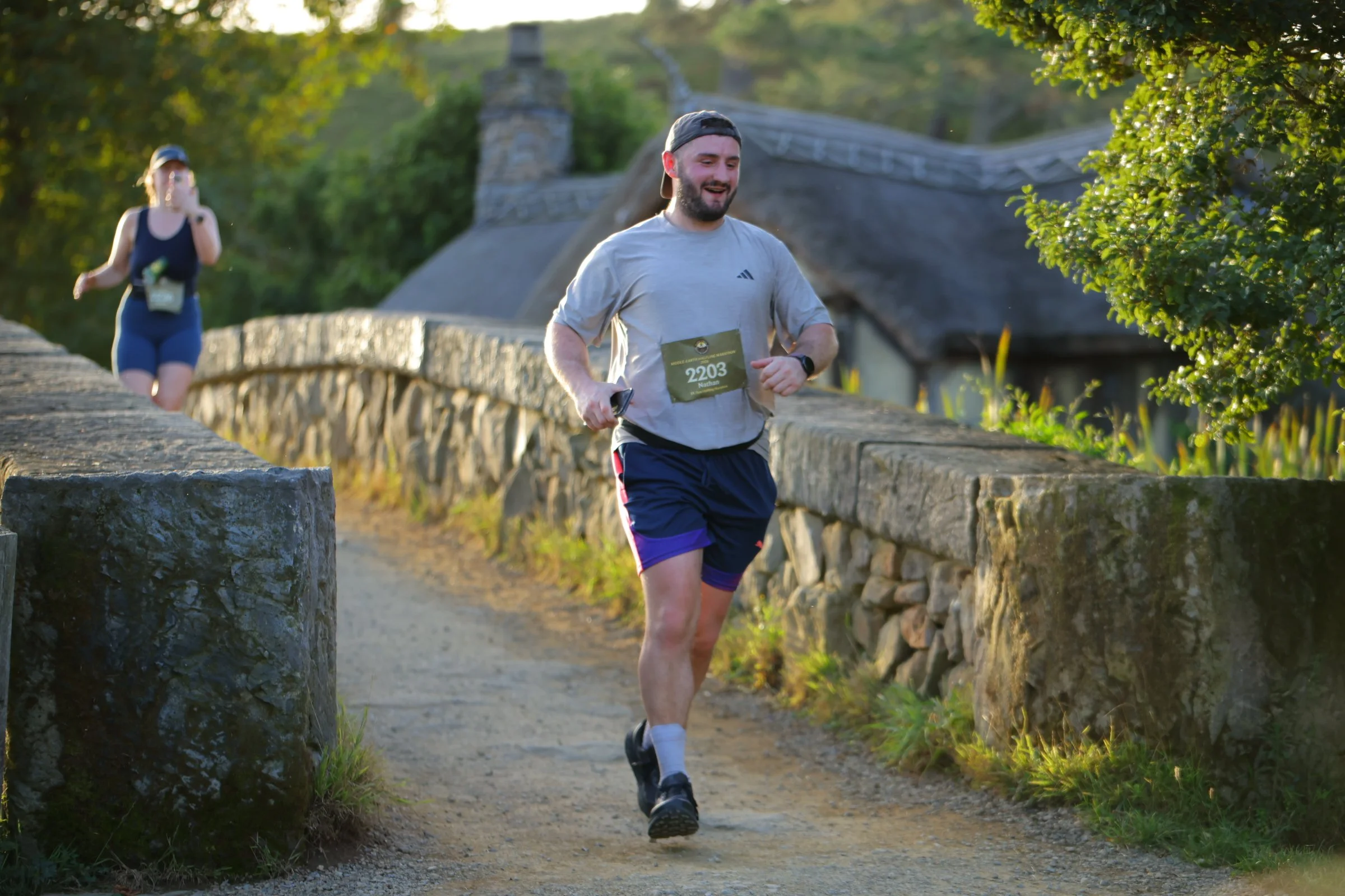 A man with a beard, wearing a gray t-shirt, black shorts with purple accents, black running shoes, a watch, and a black cap worn backwards, is running on a trail next to a stone wall during sunset. A woman wearing a black tank top and blue shorts is 