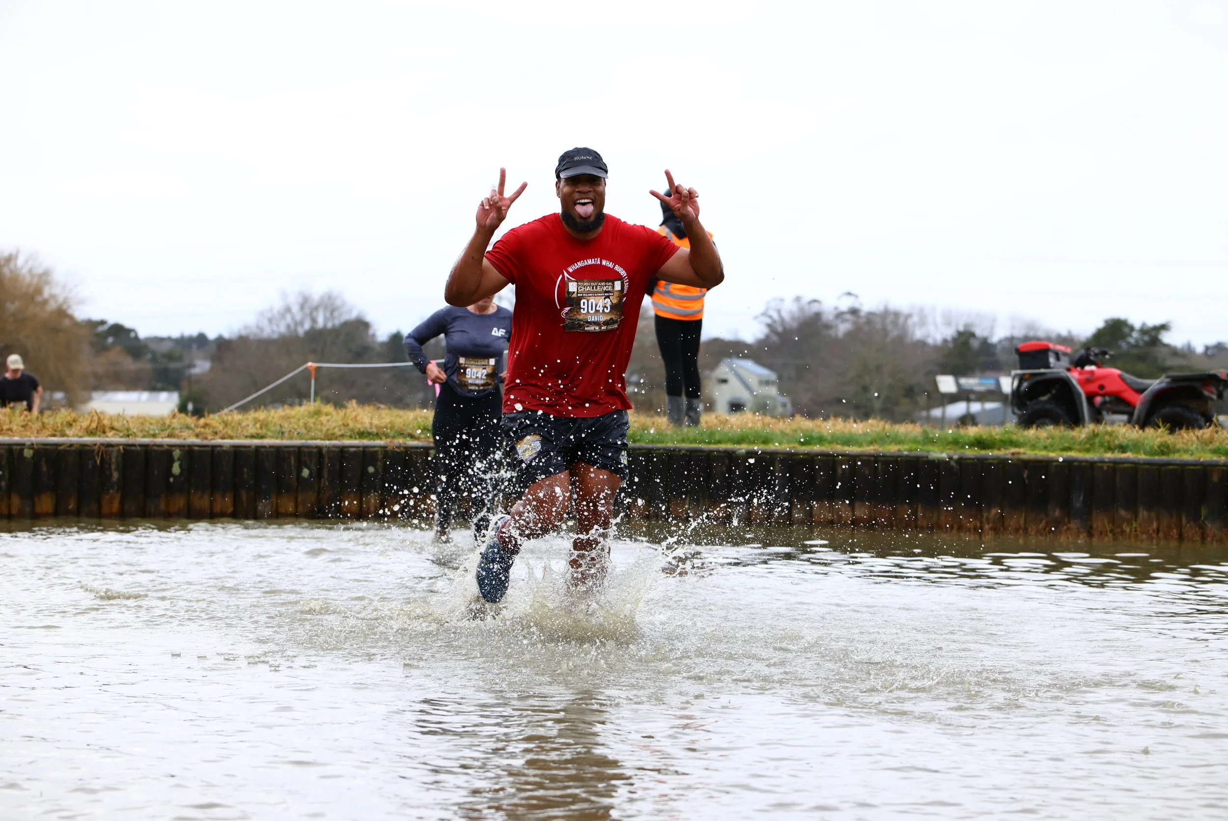 Man smiling and making peace signs while running through water during a race, with other runners and a red ATV in the background.