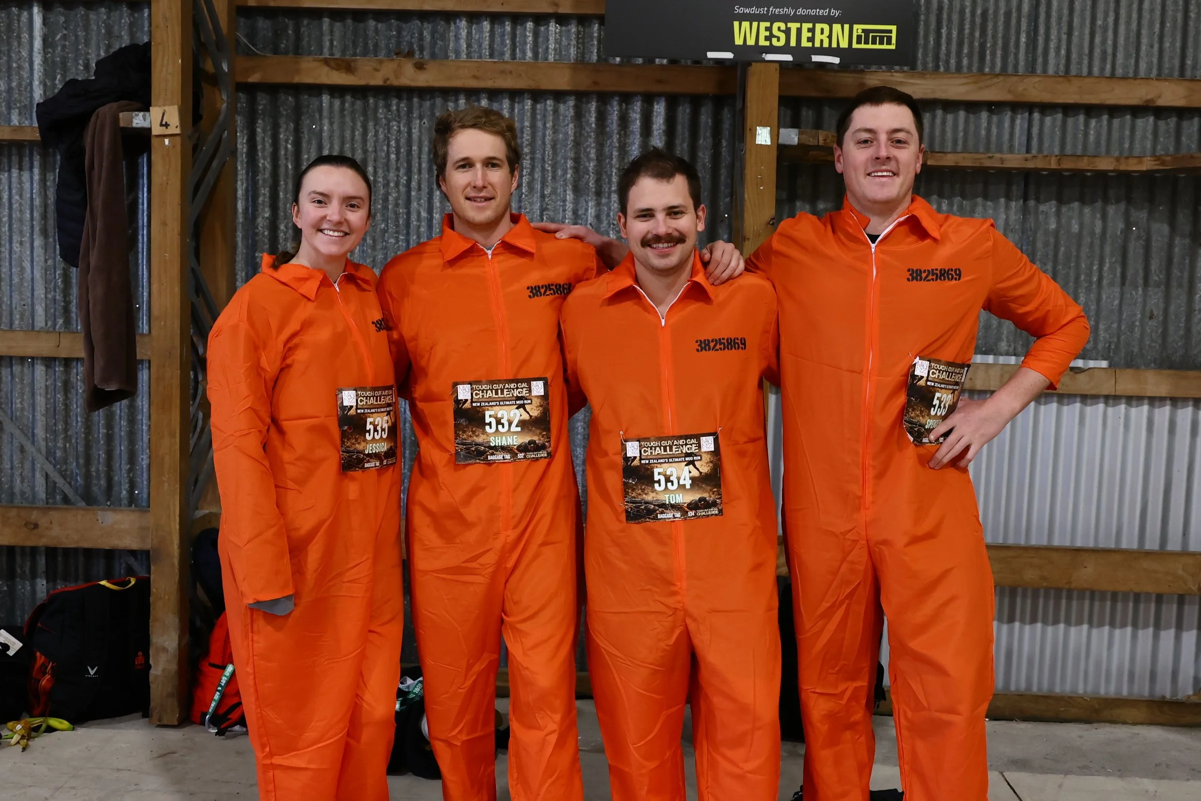 Four people dressed in orange jumpsuits with race badges on their chests are standing side by side and smiling in a rustic indoor location with corrugated metal walls. They are participating in a challenge event, as indicated by the badges.