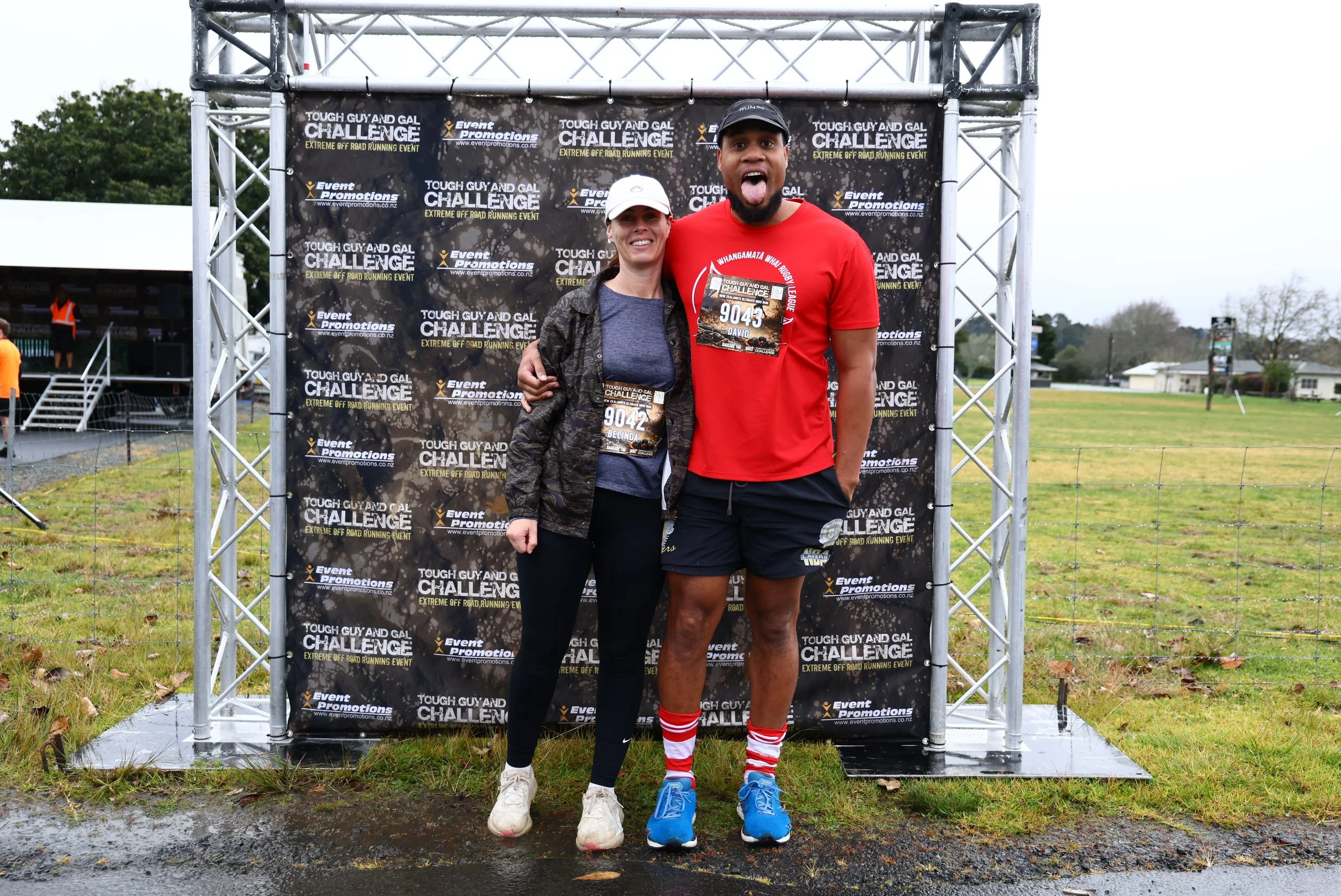 Two runners standing together in front of a black backdrop at the Tough Guy and Gal Challenge event, with their arms around each other. The woman is smiling, wearing a white cap, a gray shirt under a black jacket, black leggings, and white shoes. The