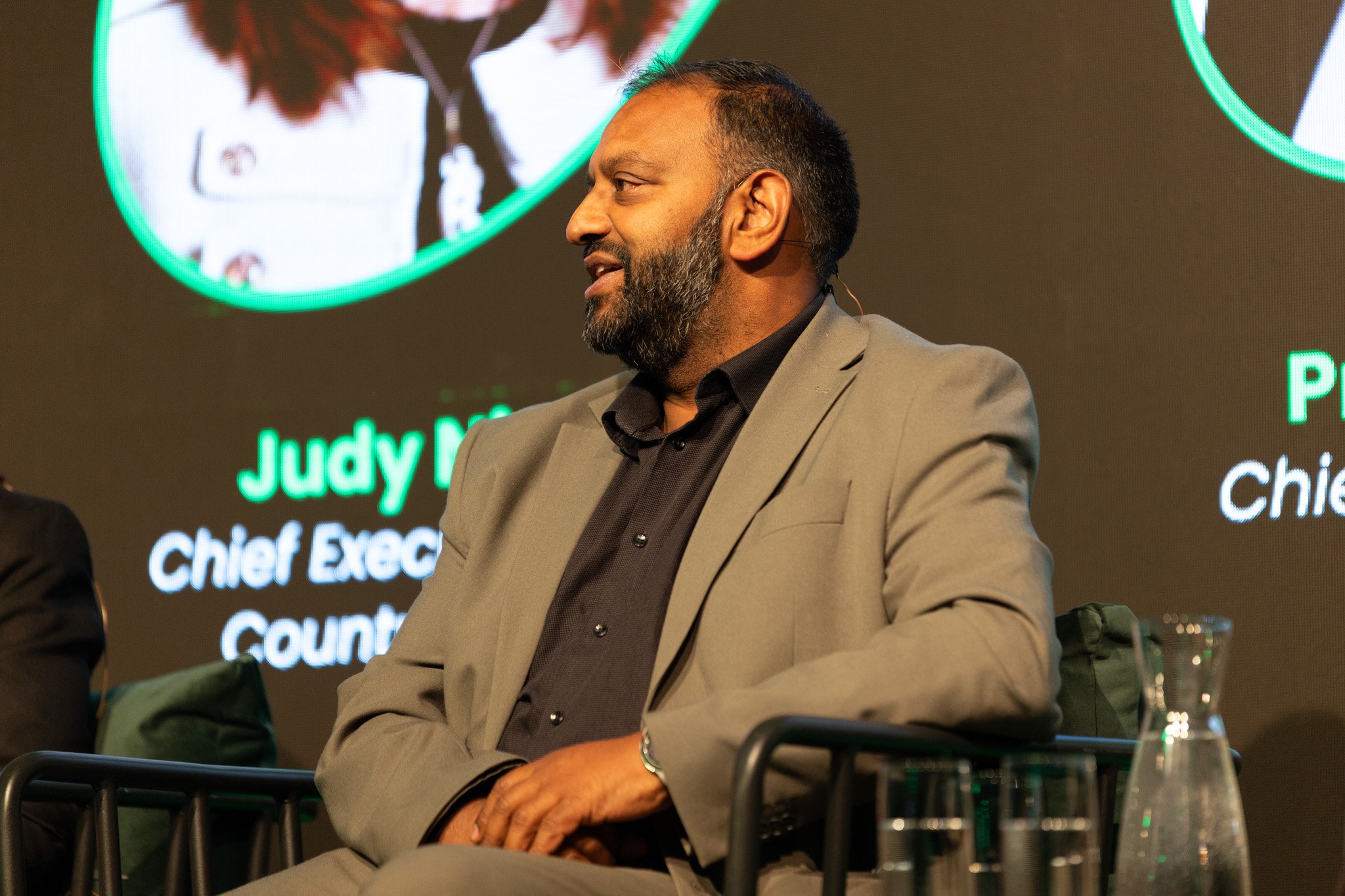 A man with a beard and dark hair, wearing a gray blazer and black shirt, is sitting at a panel or conference. There is a large screen behind him displaying text and graphics related to Judy, a chief executive, and county figures. The man is looking t