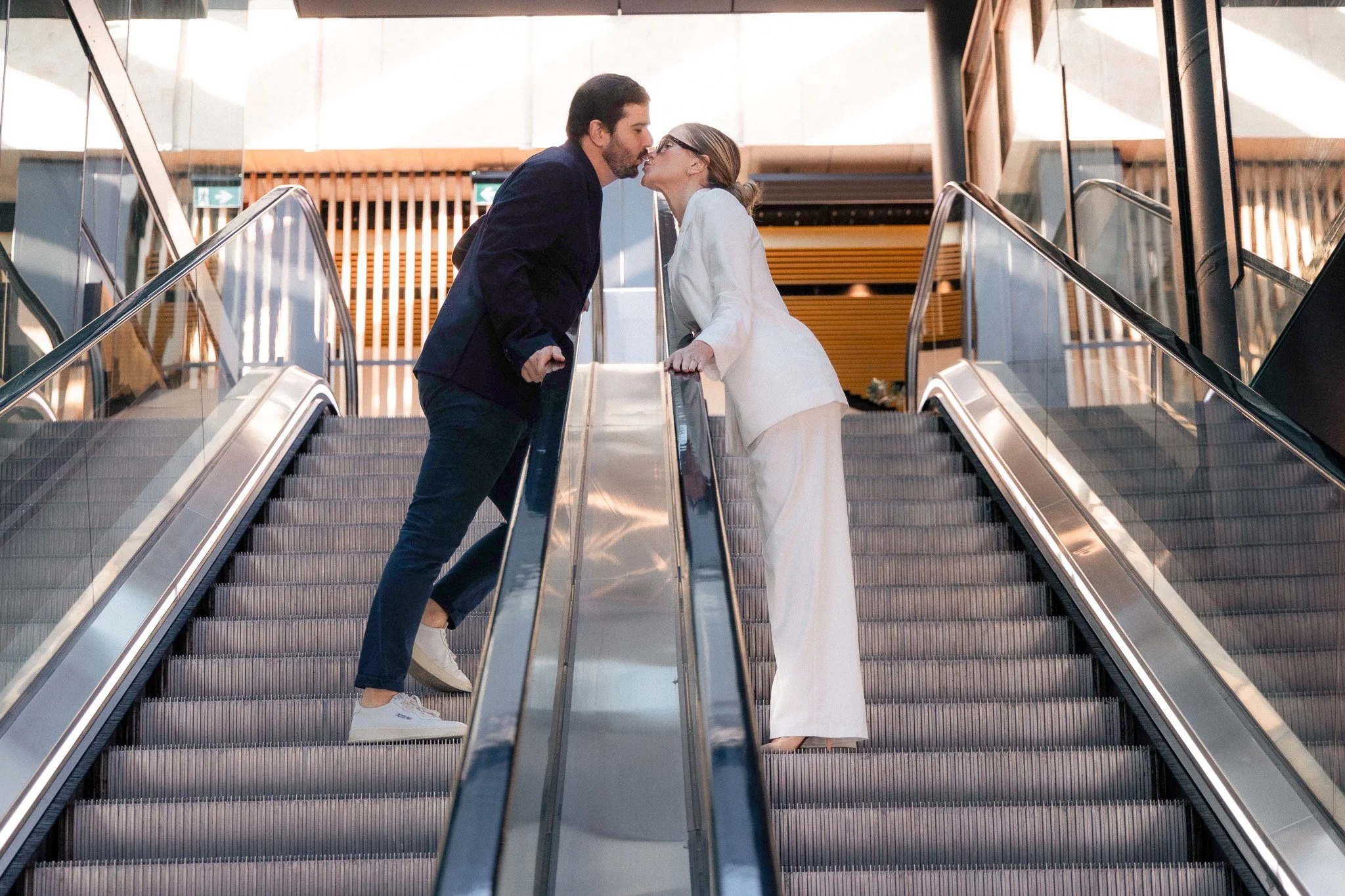 A man and woman on an escalator in a modern building leaning in for a kiss.