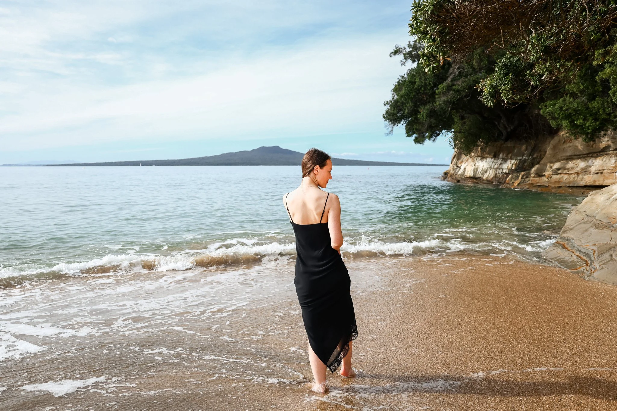 A woman in a black dress walking along the sandy shore of a body of water, with waves gently hitting the beach. There are trees and rocky cliffs on the right side and an island with a mountain in the background. The sky is partly cloudy.