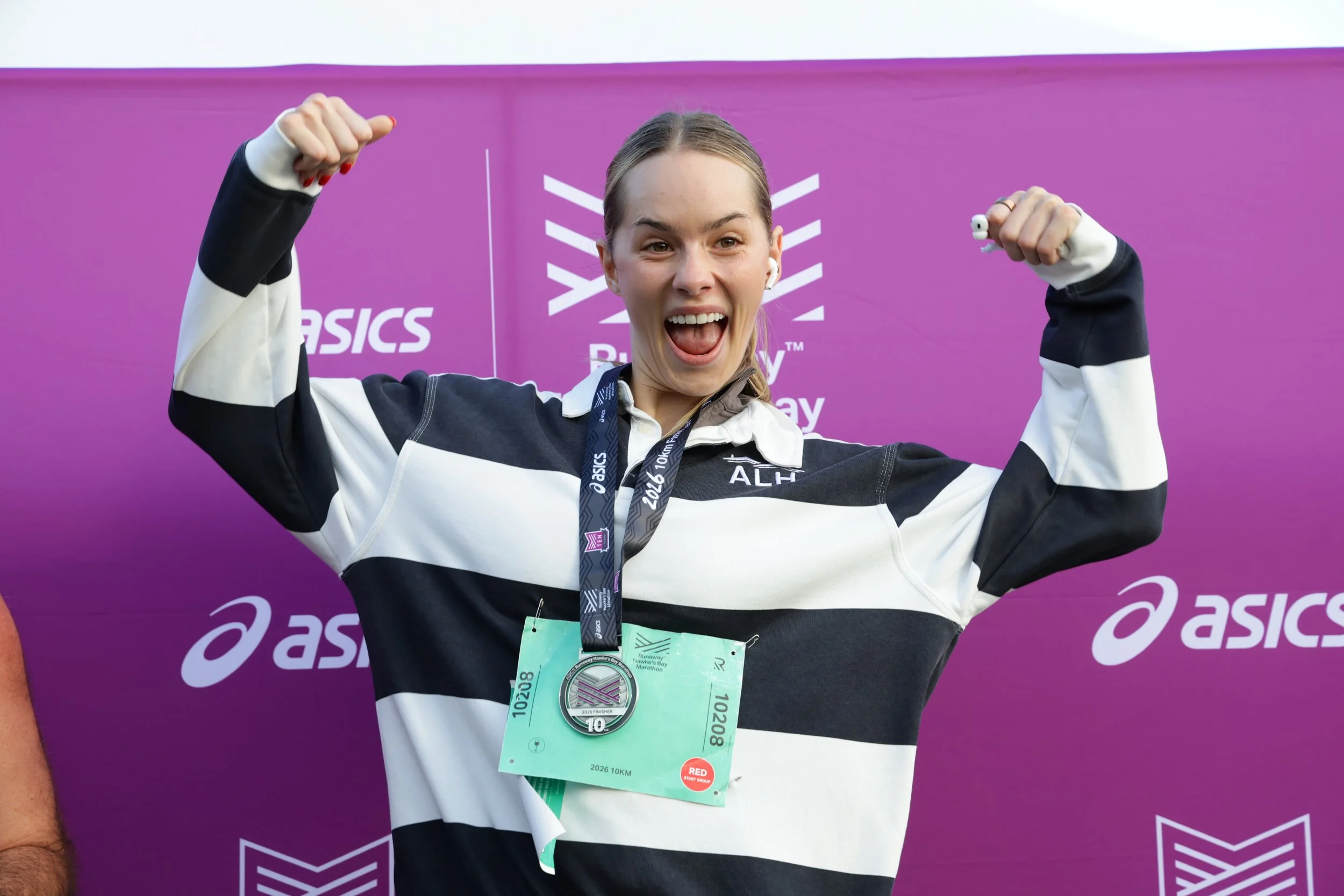 A woman celebrating after finishing a race, wearing a black and white striped shirt, with a finisher’s medal around her neck, and a race bib on her chest, in front of a purple backdrop with branding.
