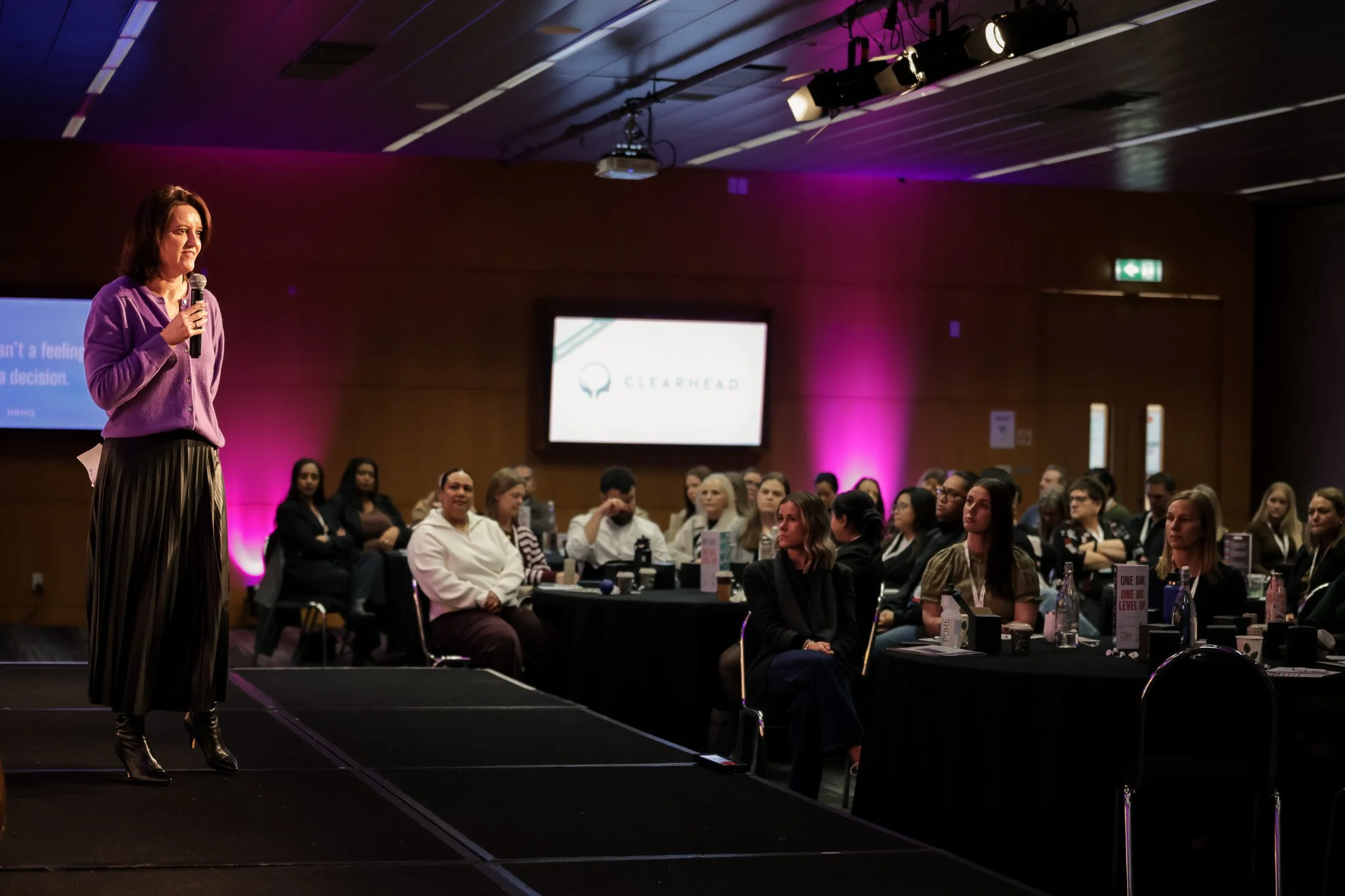 A woman in a purple sweater and striped pants stands on a stage with a microphone, addressing an audience of diverse adults seated at tables in a dimly lit conference room with purple lighting and two large screens on the wall.