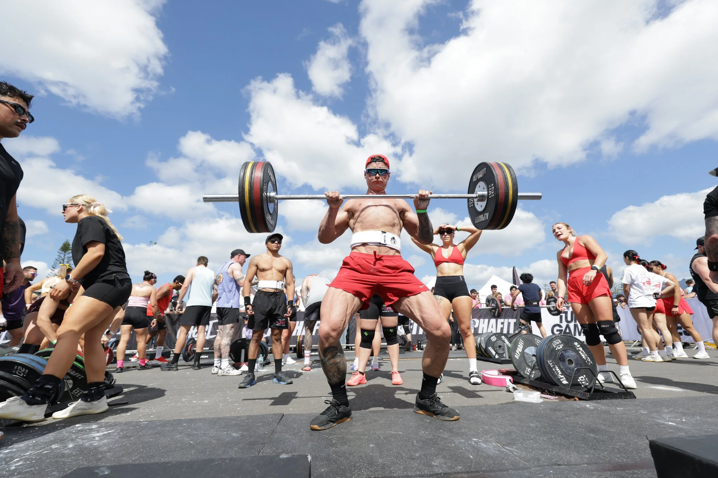 Man lifting a barbell at outdoor fitness event with people around, under blue sky with clouds.