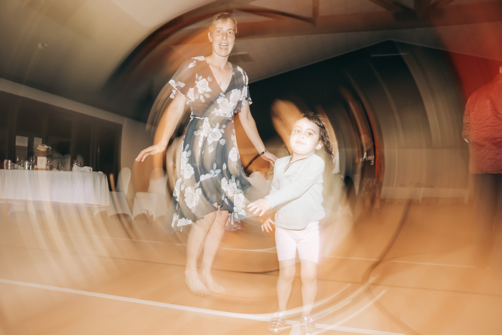 A woman in a floral dress and a young girl in a white jacket are dancing together in a room with a wooden ceiling. The photo has a swirling motion effect around the edges.