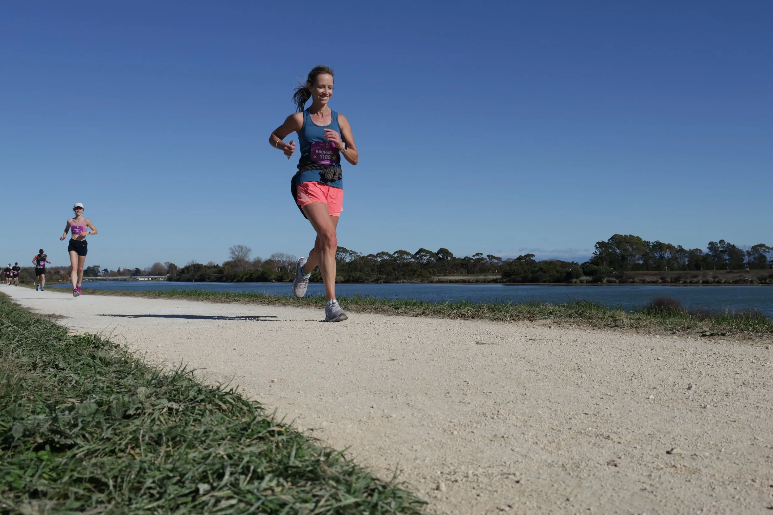 A woman running on a dirt path next to a body of water under a clear blue sky, wearing a blue tank top and pink shorts, with other runners visible in the background.