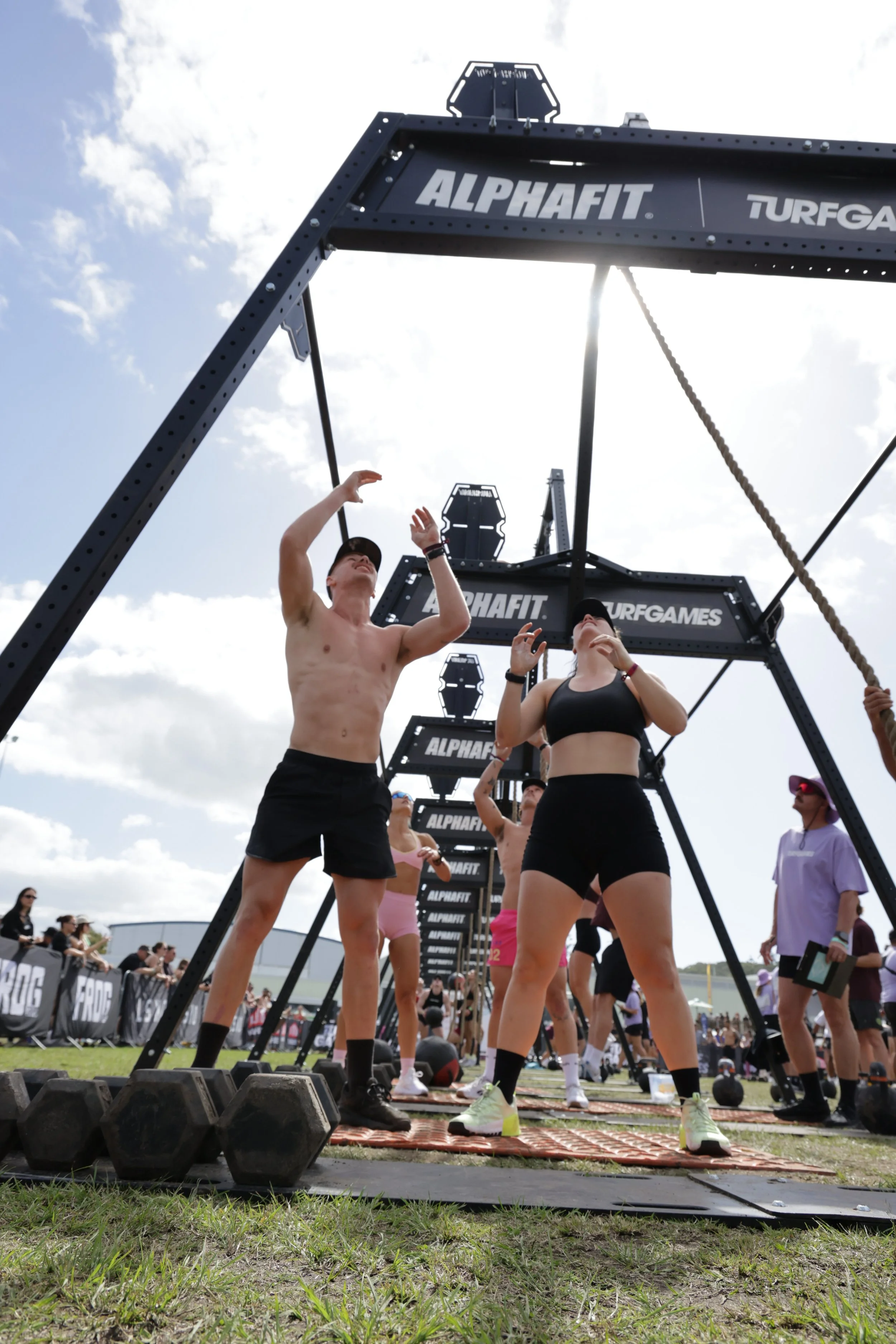 Participants preparing for a CrossFit competition outdoors, with fitness equipment and a large ATLAS-style rig structure branded with 'ALPHAFIT' and 'TURFGA'. People are stretching and warming up on mats and ground, under partly cloudy skies.