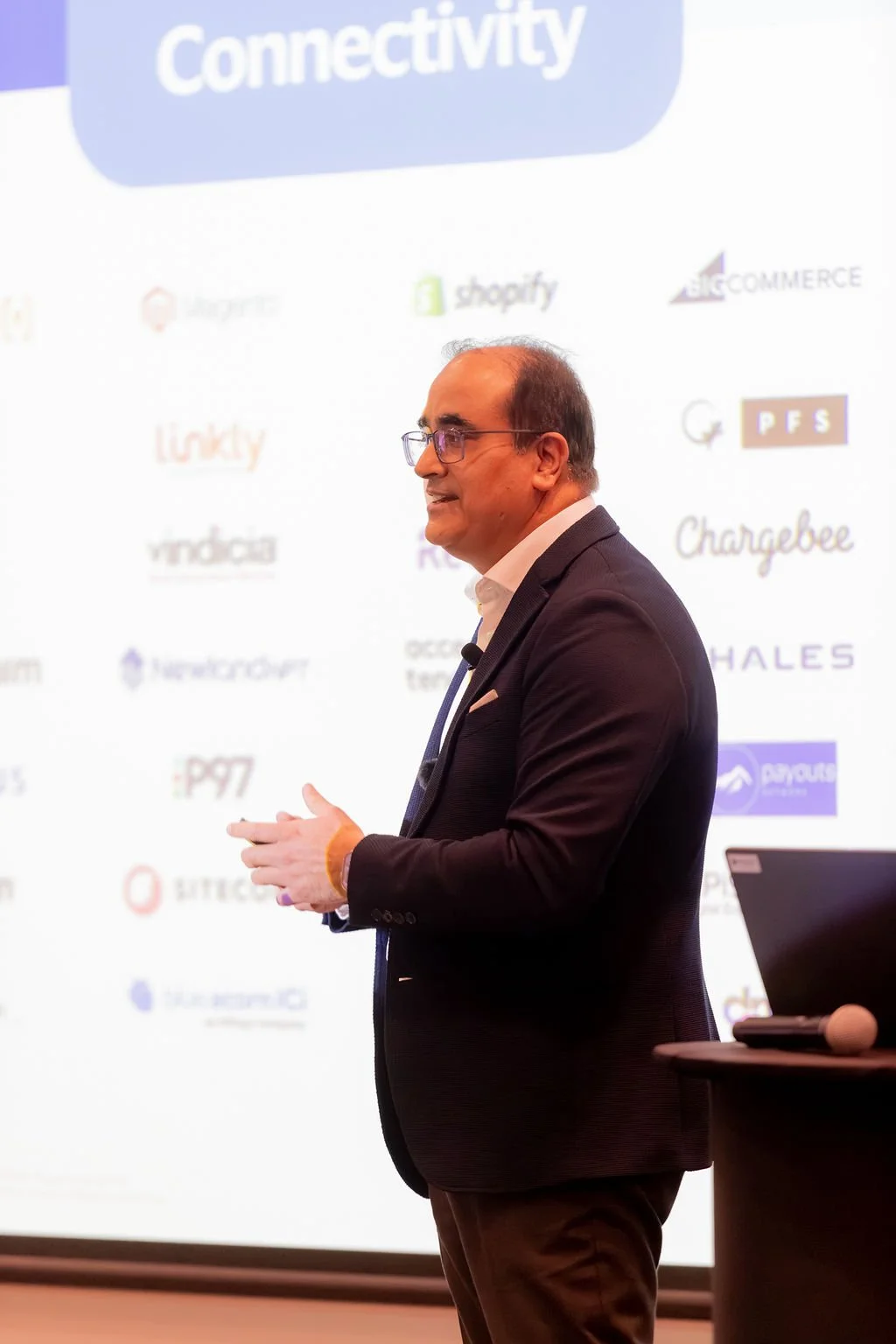 A man in a dark suit with glasses, standing in front of a presentation screen, speaking or presenting at a conference.
