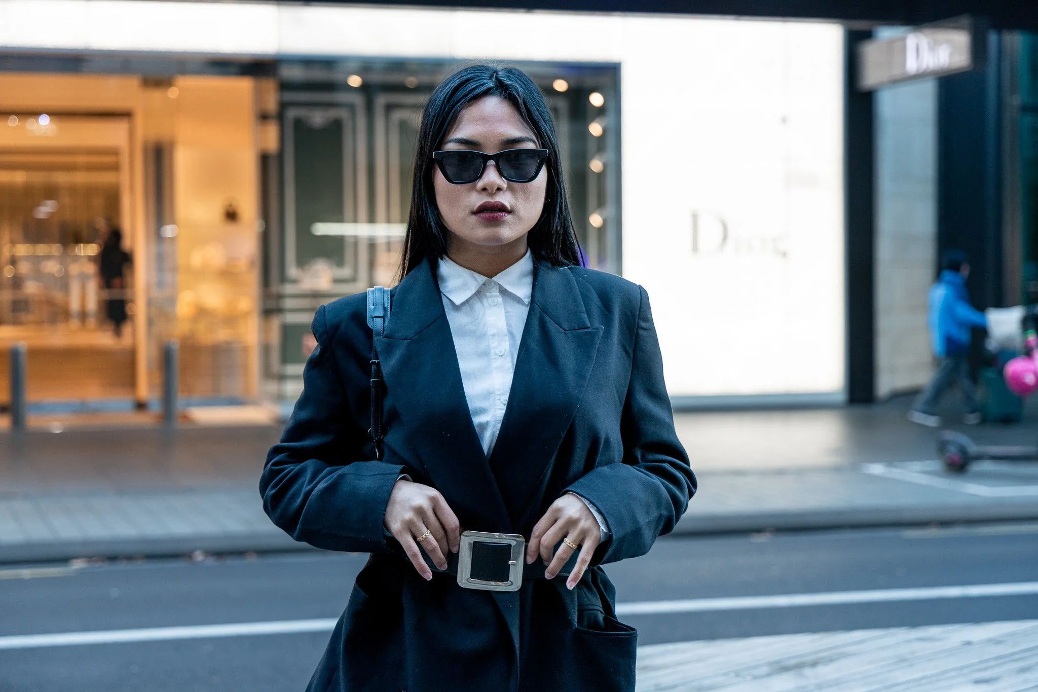 A woman with black hair wearing dark sunglasses, a white collared shirt, and a dark blazer stands outside on a city sidewalk, holding a smartphone.