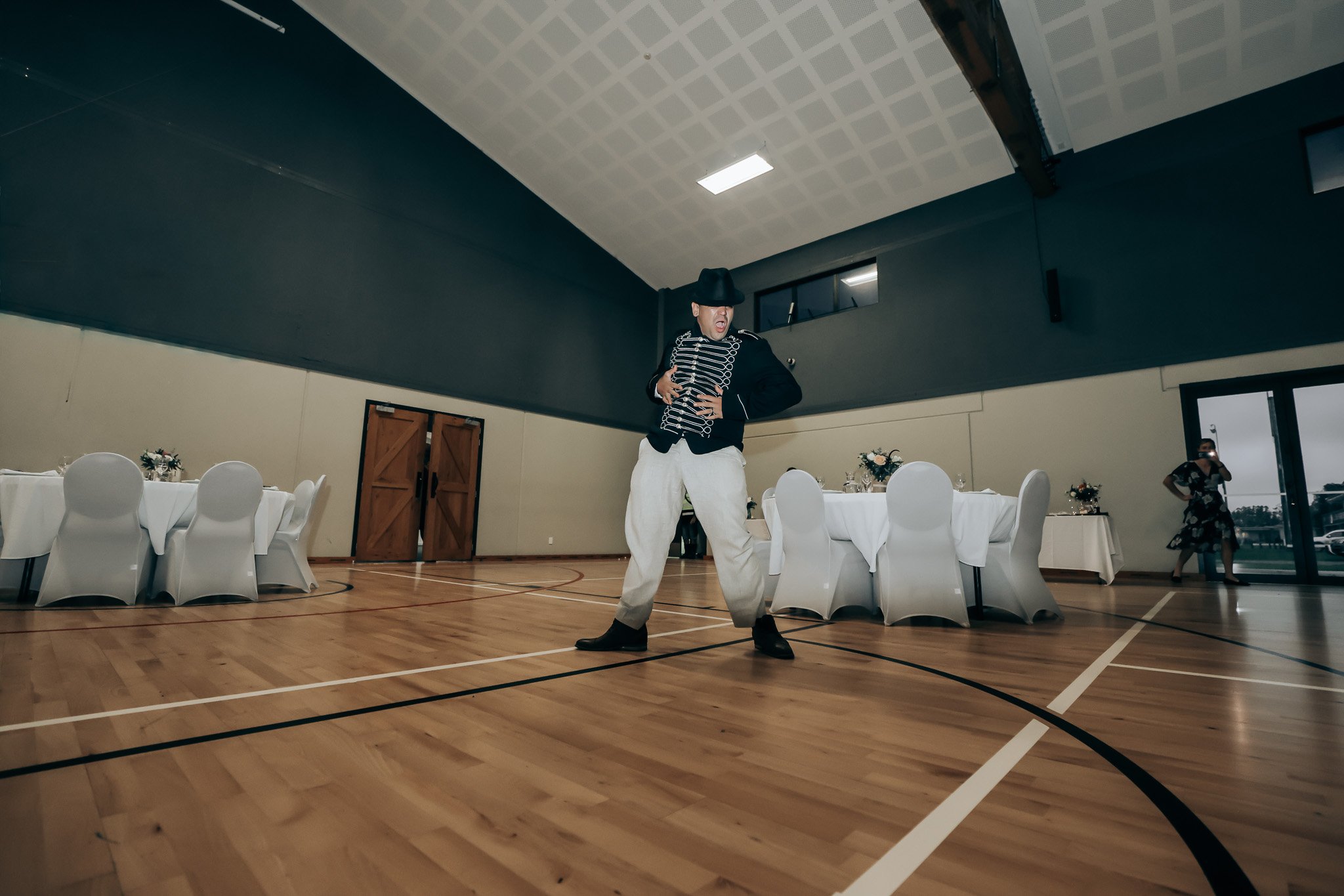 A man in a black hat and military-style jacket dancing in an indoor gymnasium, with round tables and chairs in the background.
