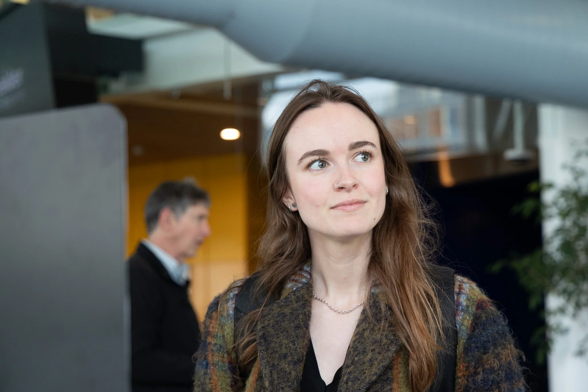 A young woman with long brown hair and blue eyes looking to the side, wearing a patterned jacket and a necklace, in an indoor setting with blurred background of a man and other people.