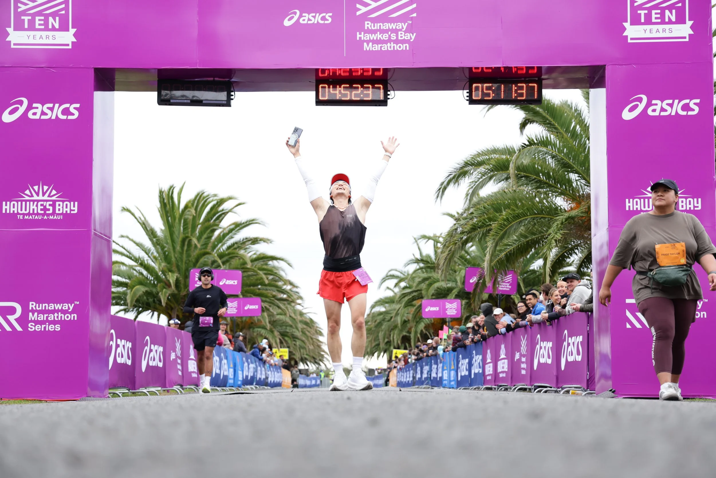 A woman running across the finish line at a marathon event, celebrating with arms raised. The event is the Hawke's Bay Marathon, sponsored by ASICS, with crowd support on either side and palm trees in the background.