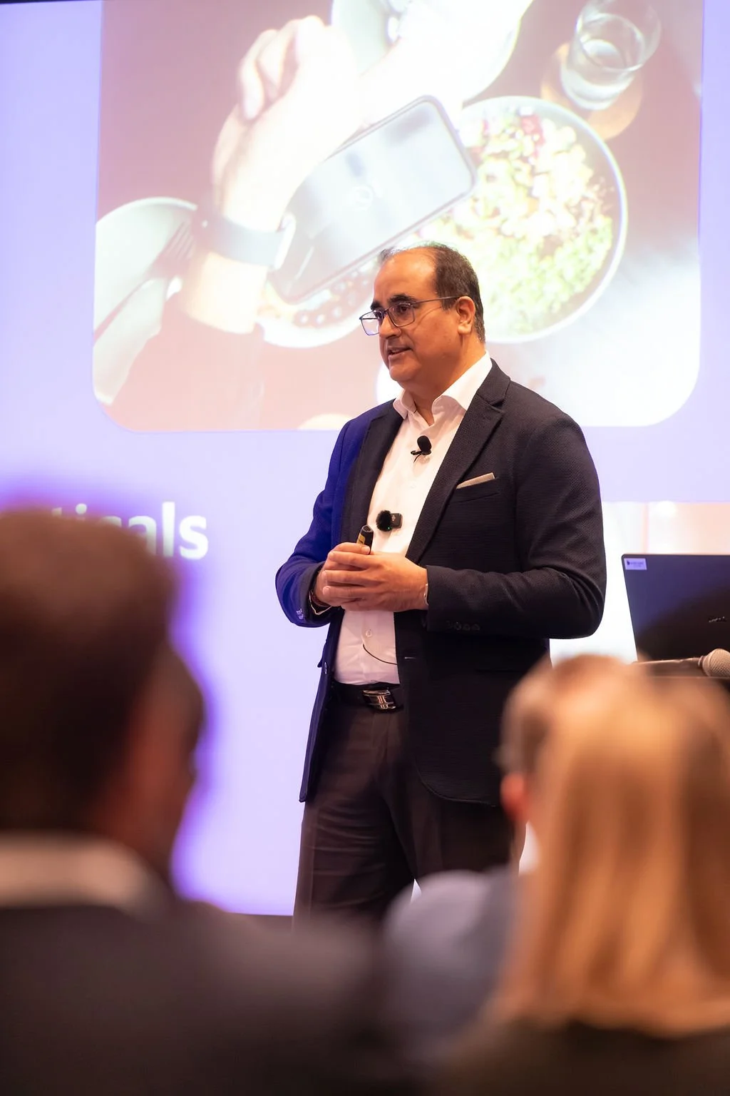 A man in a dark suit and glasses giving a presentation in front of a screen showing a food photo, with an audience in the foreground.