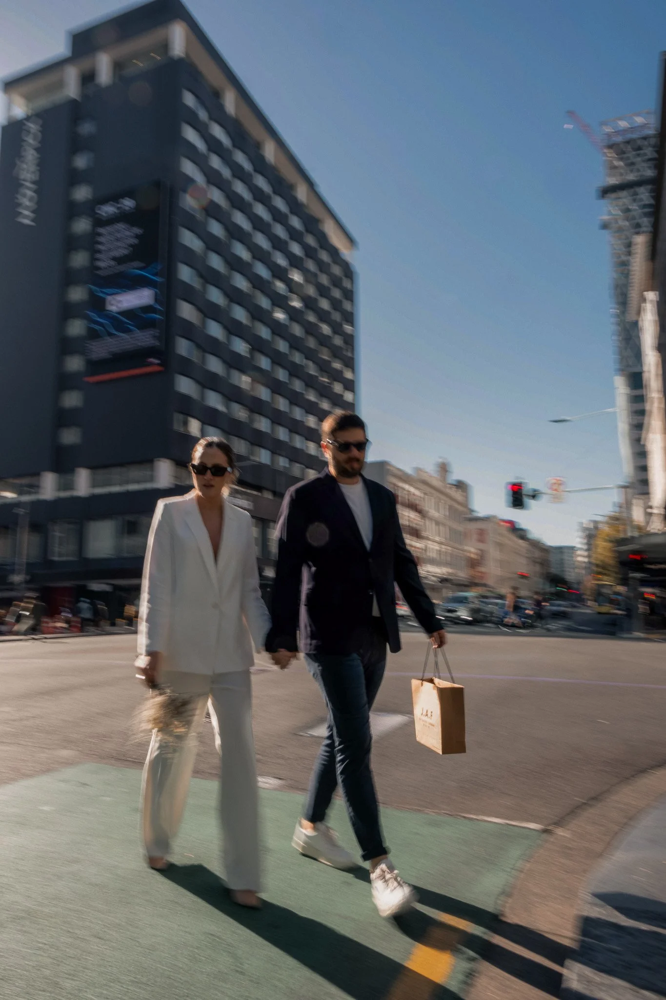A woman in a white suit and a man in a dark suit walk hand in hand in an urban setting, holding shopping bags. The background features modern high-rise buildings, a clear blue sky, and traffic lights.