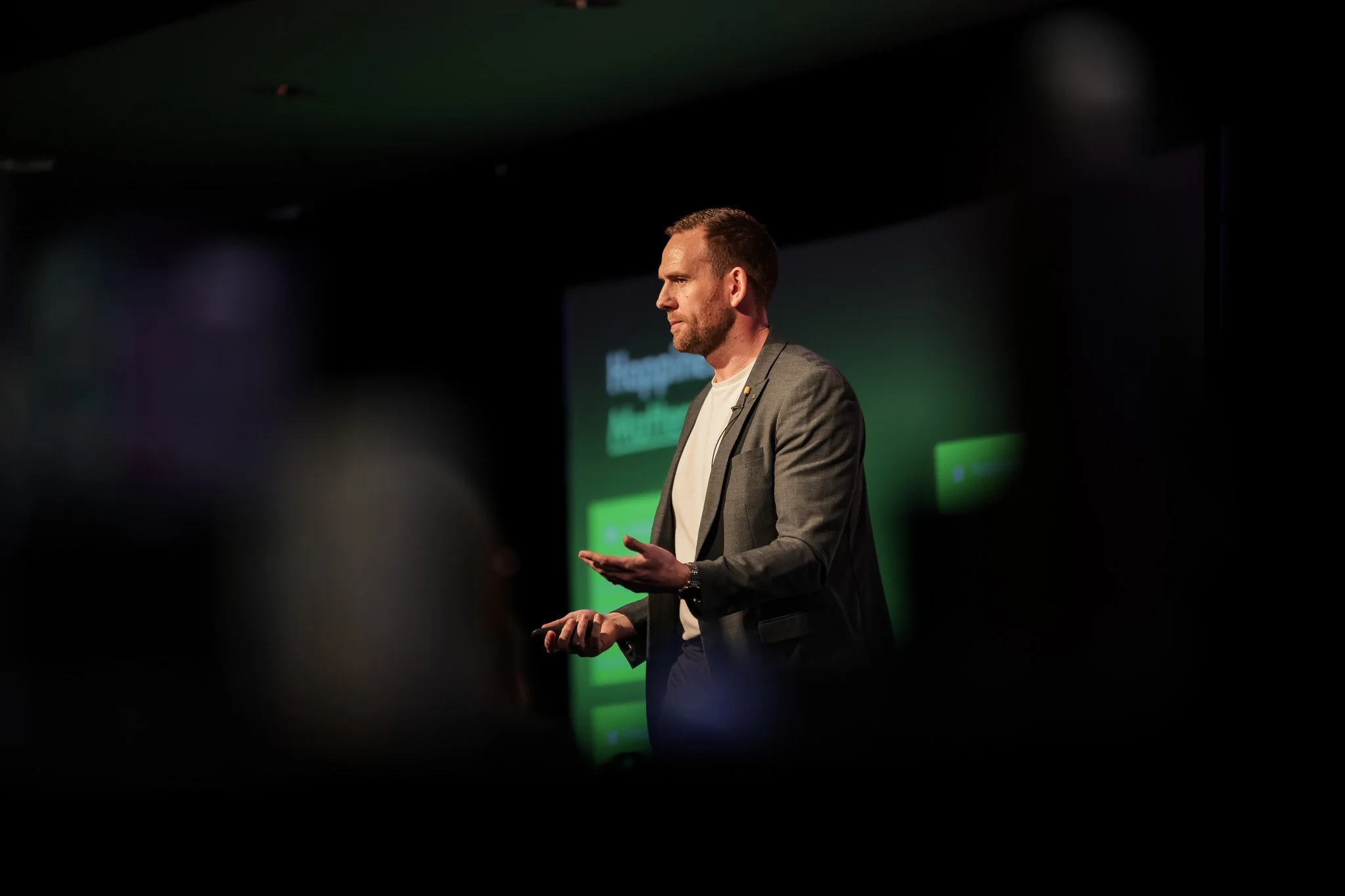 A man in a gray blazer and white shirt giving a presentation on stage with a green background.