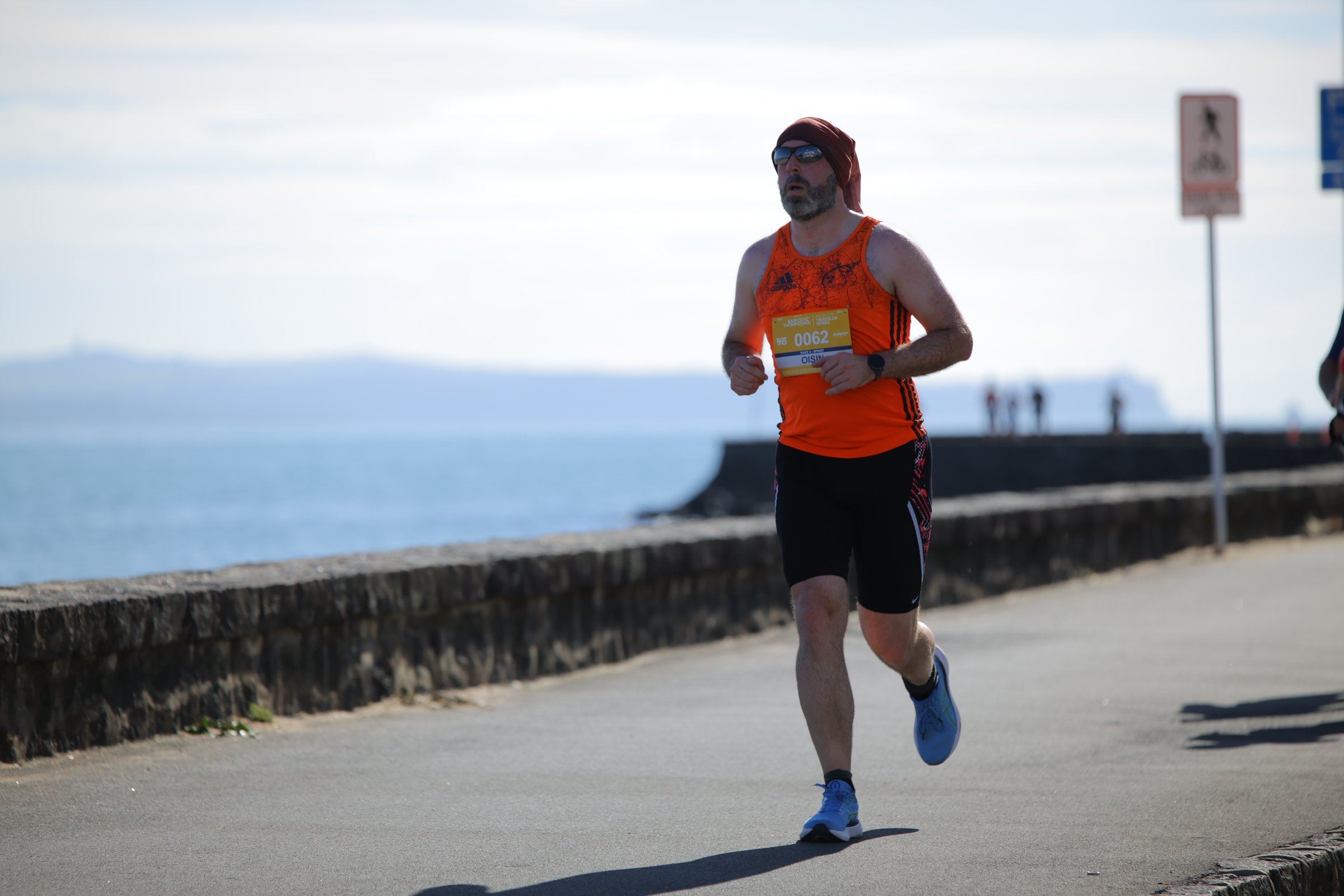 A man running along a waterfront promenade wearing a red tank top, black shorts, sunglasses, and a head wrap. He has a race bib number 0062 and appears focused on his run.