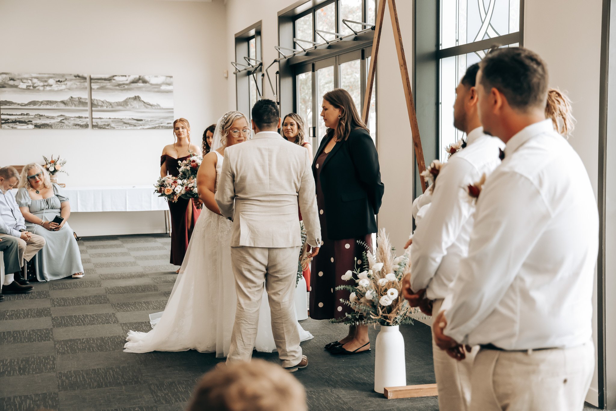 A wedding ceremony with the bride and groom facing each other, surrounded by attendants and guests in a modern indoor setting.